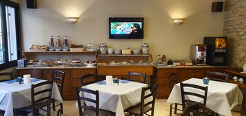 Restaurante com mesas montadas para refeições, uma televisão na parede e diversos dispensadores de comida e bebida no balcão.