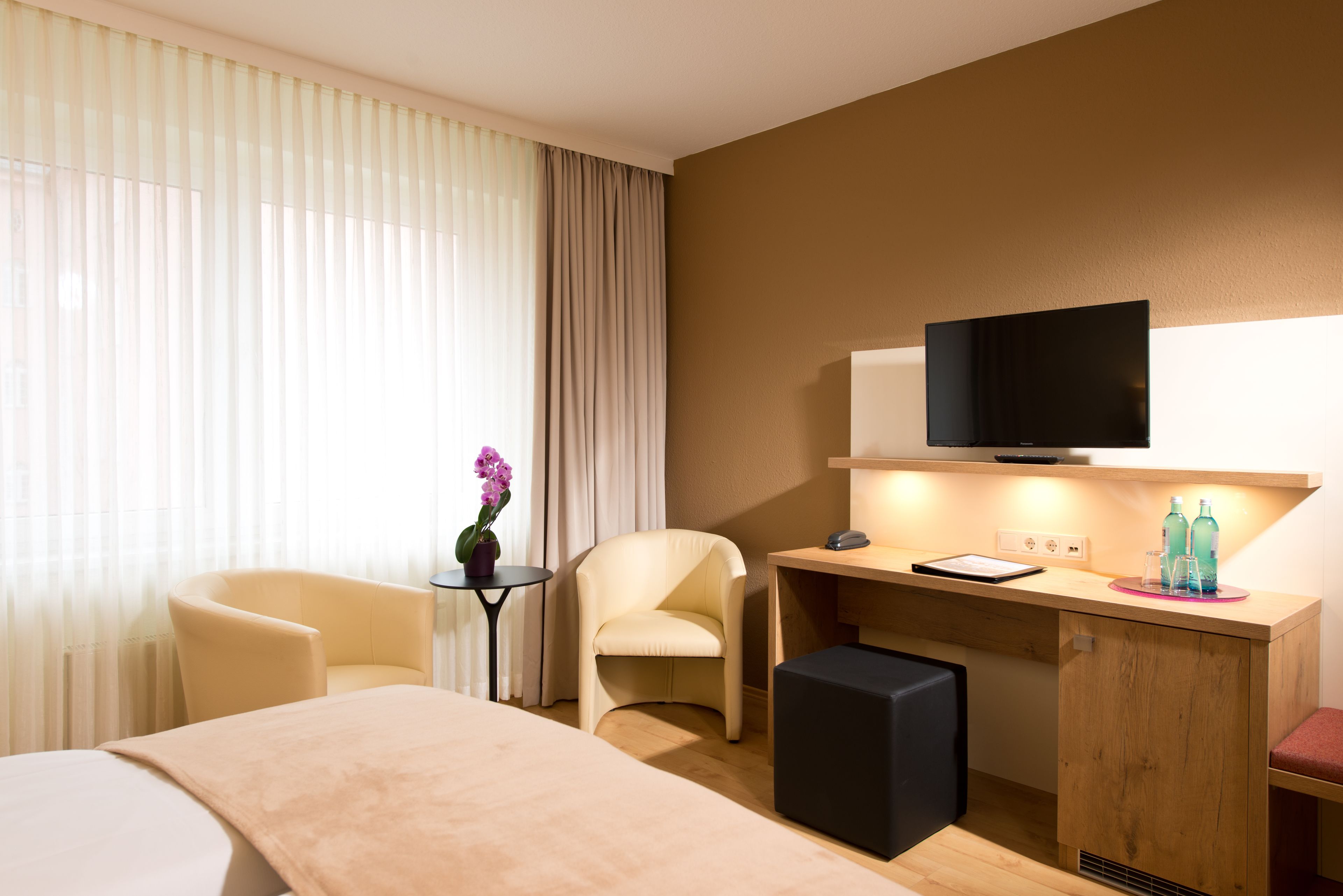 Habitación de hotel con cama, escritorio con televisor, una silla, una mesita con un florero y una ventana con cortinas.