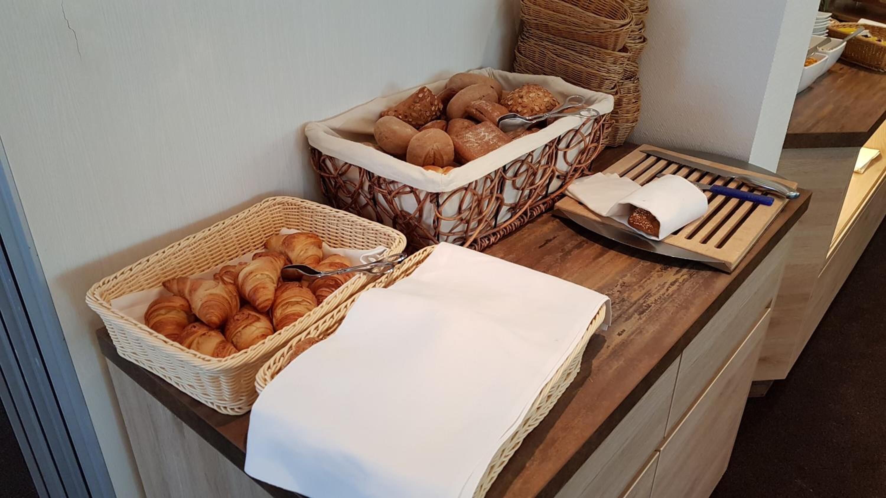 Buffet de café da manhã com croissants, pães e uma bandeja com um cachorro-quente e utensílios.