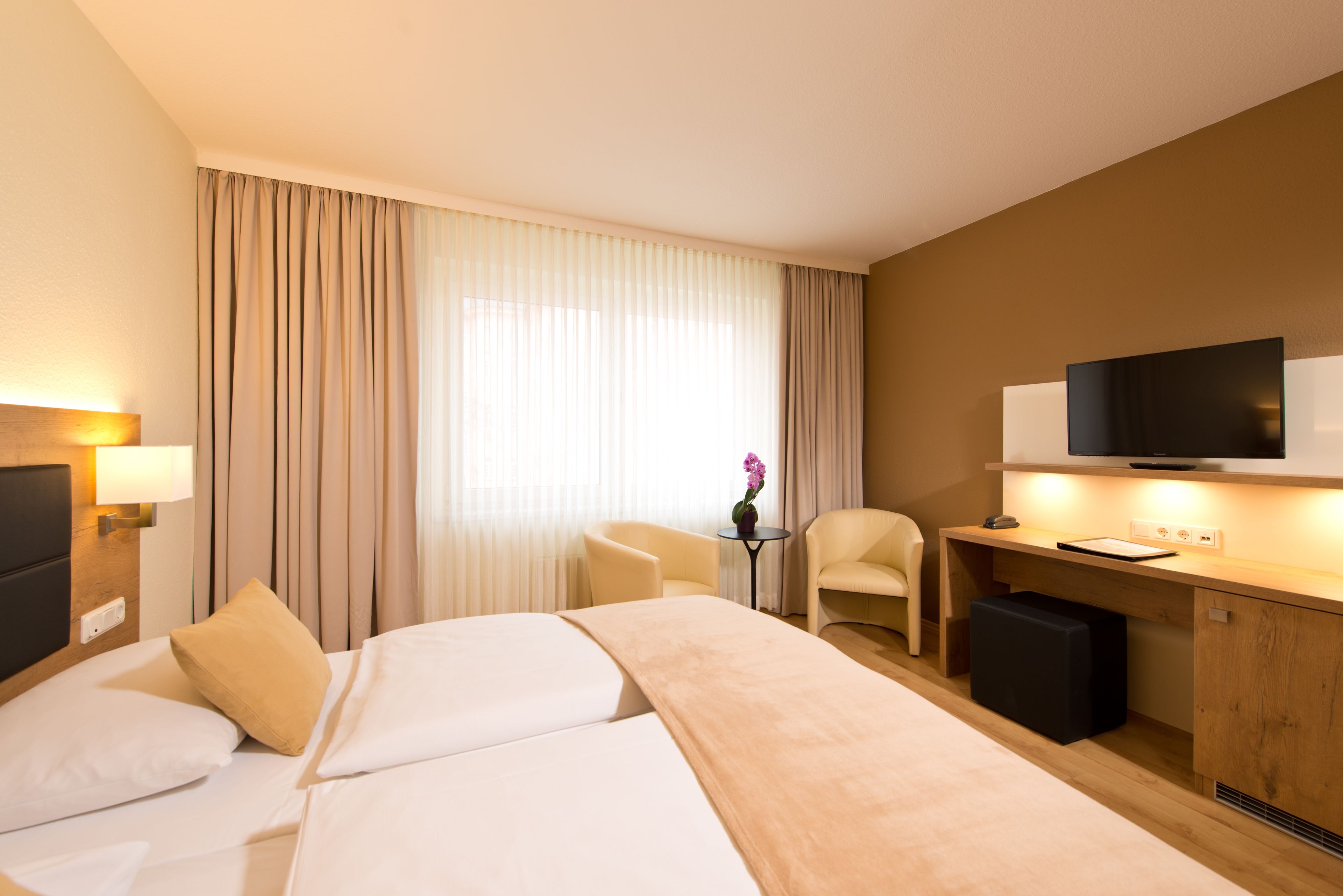 A hotel room with a bed, a desk, a chair, a television, and a small table with a flower.