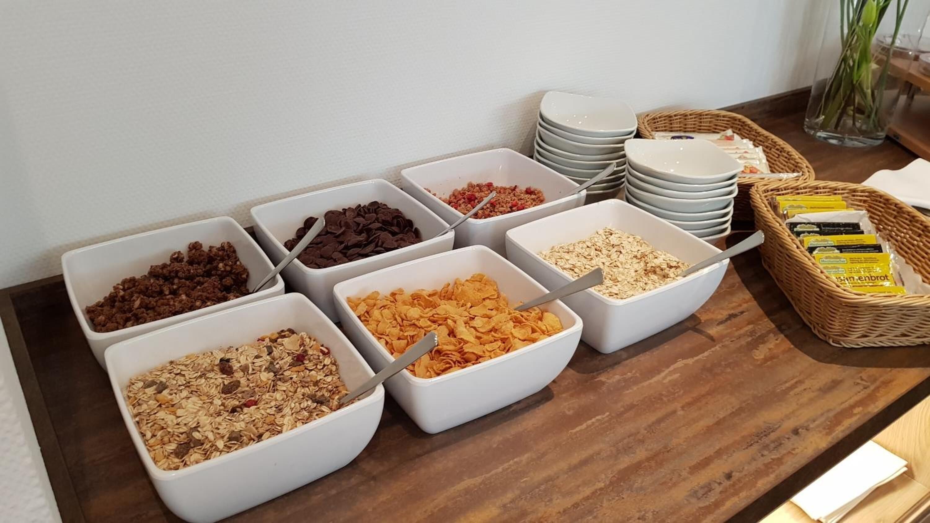 Un bufé con diversos alimentos en tazones blancos, incluyendo cereales, chispas de chocolate y granola.