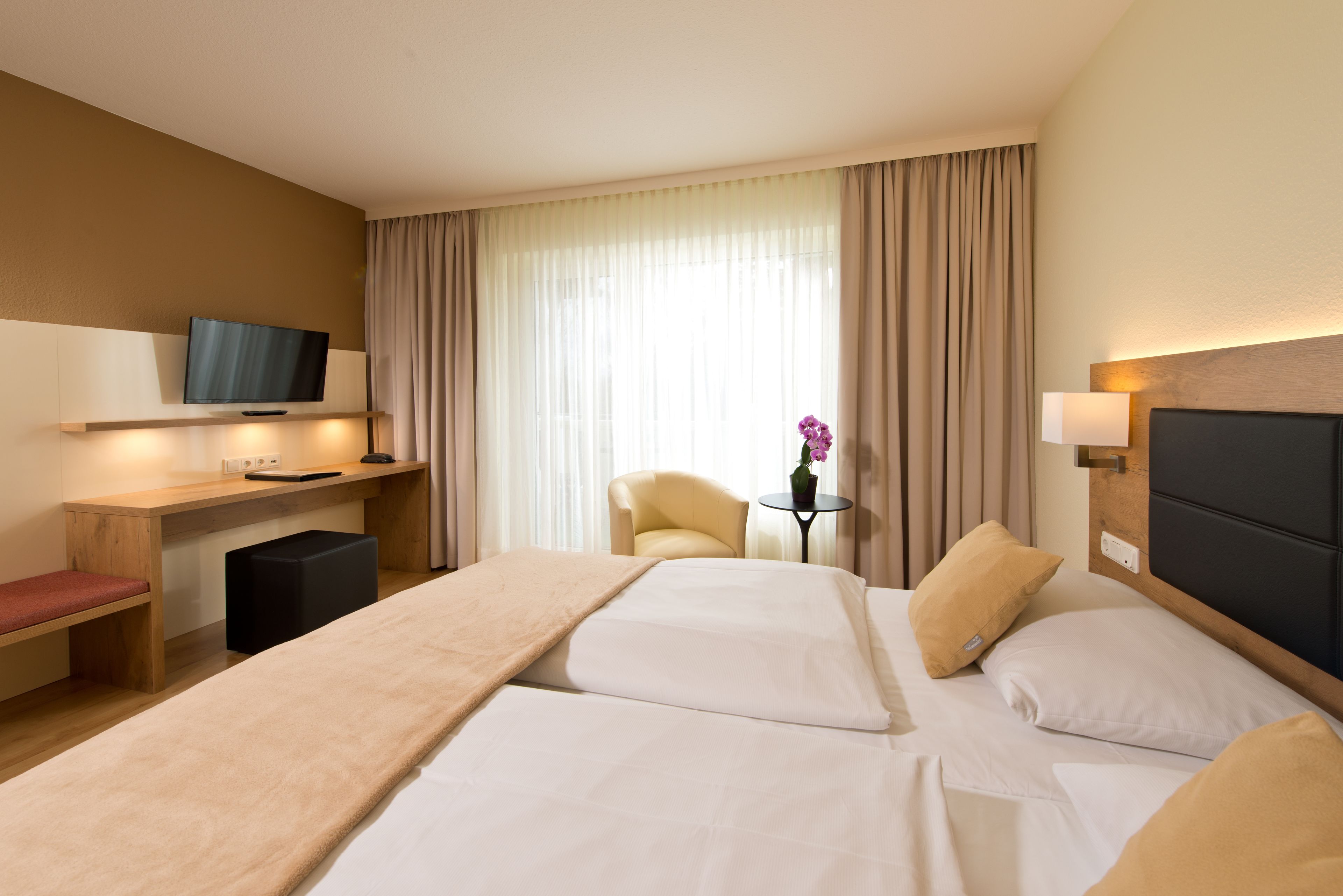 Quarto de hotel com uma cama grande, uma escrivaninha com televisão, uma cadeira e uma pequena mesa com um vaso de flores.