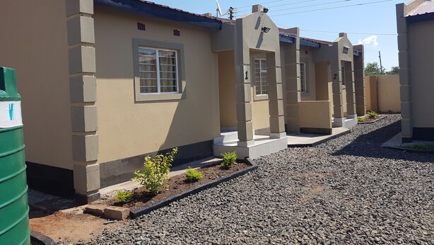 A row of beige houses with numbered windows, a green trash bin, and gravel pathways. - Sumbulwa Apartments (Livingstone)