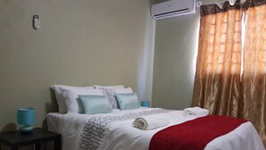 A neatly made bed with a white and red blanket, a blue lamp on a side table, and a wall-mounted air conditioner. - Sumbulwa Apartments (Livingstone)