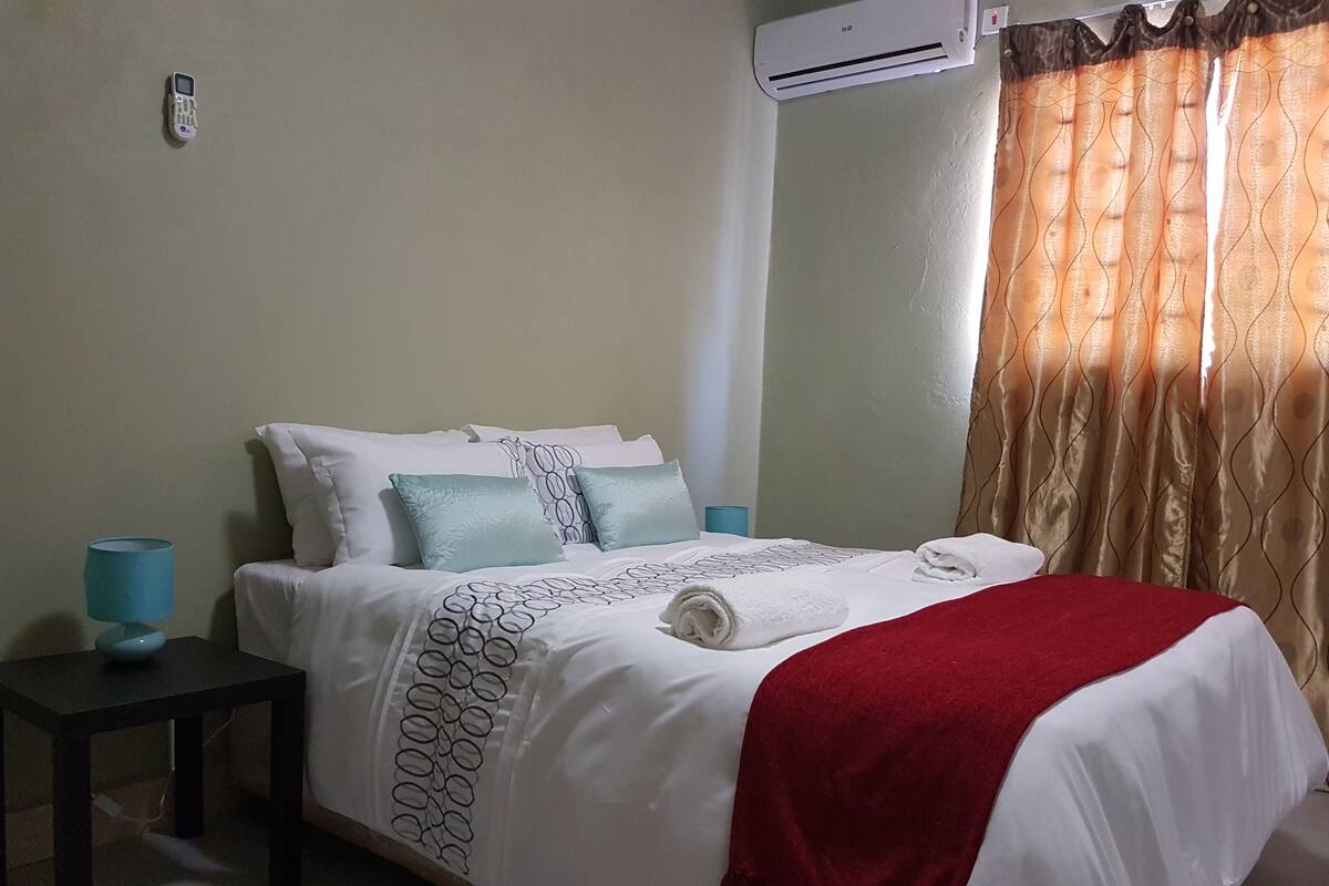 A neatly made bed with a white and red blanket, a blue lamp on a side table, and a wall-mounted air conditioner.
