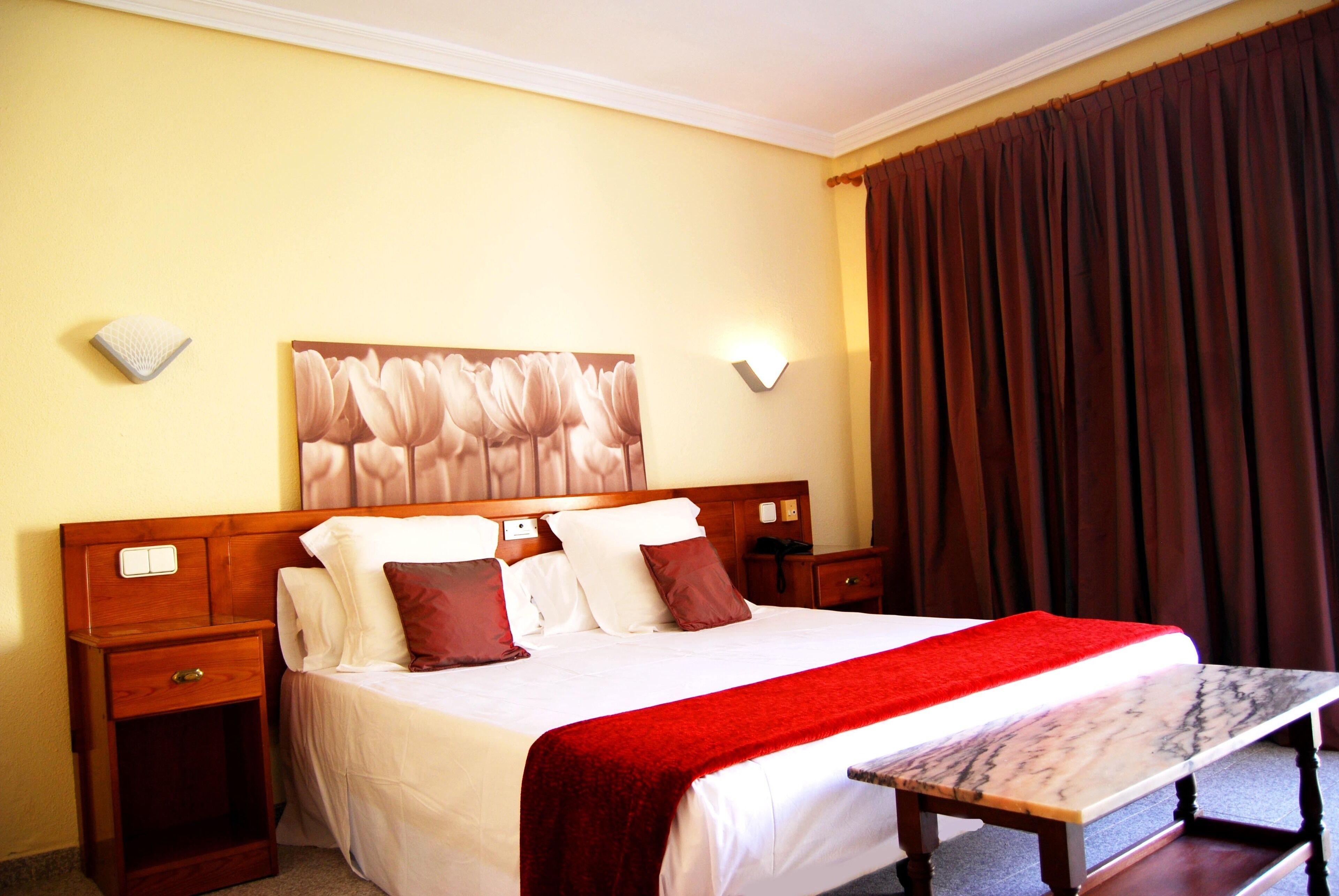 A hotel room with a large bed, wooden headboard, two bedside tables, a nightstand, a red throw blanket, a marble bedside table, and a wall-mounted light fixture.