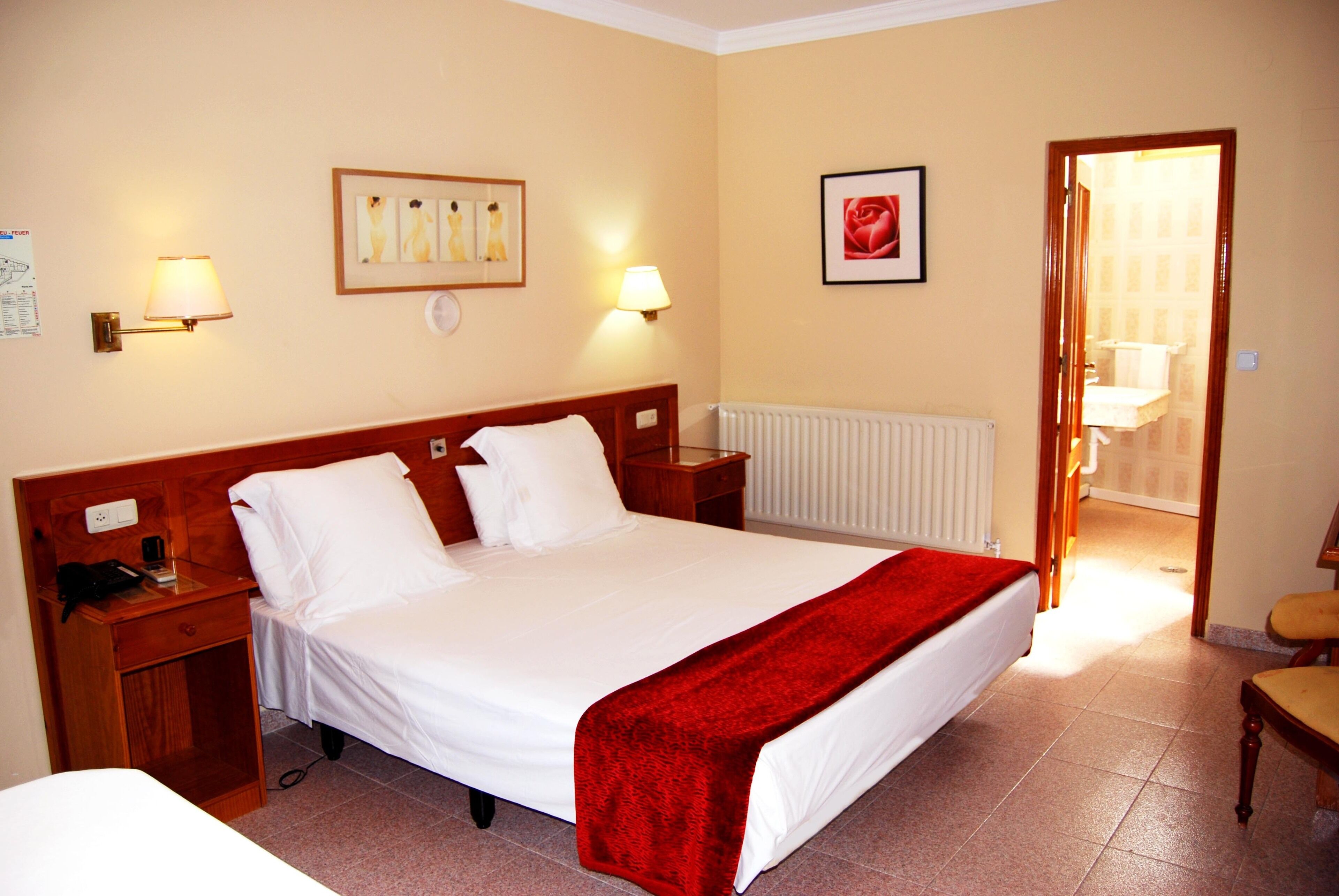 Habitación de hotel con una cama grande, dos mesitas de noche, un radiador y un edredón rojo, además de un cuadro en la pared.