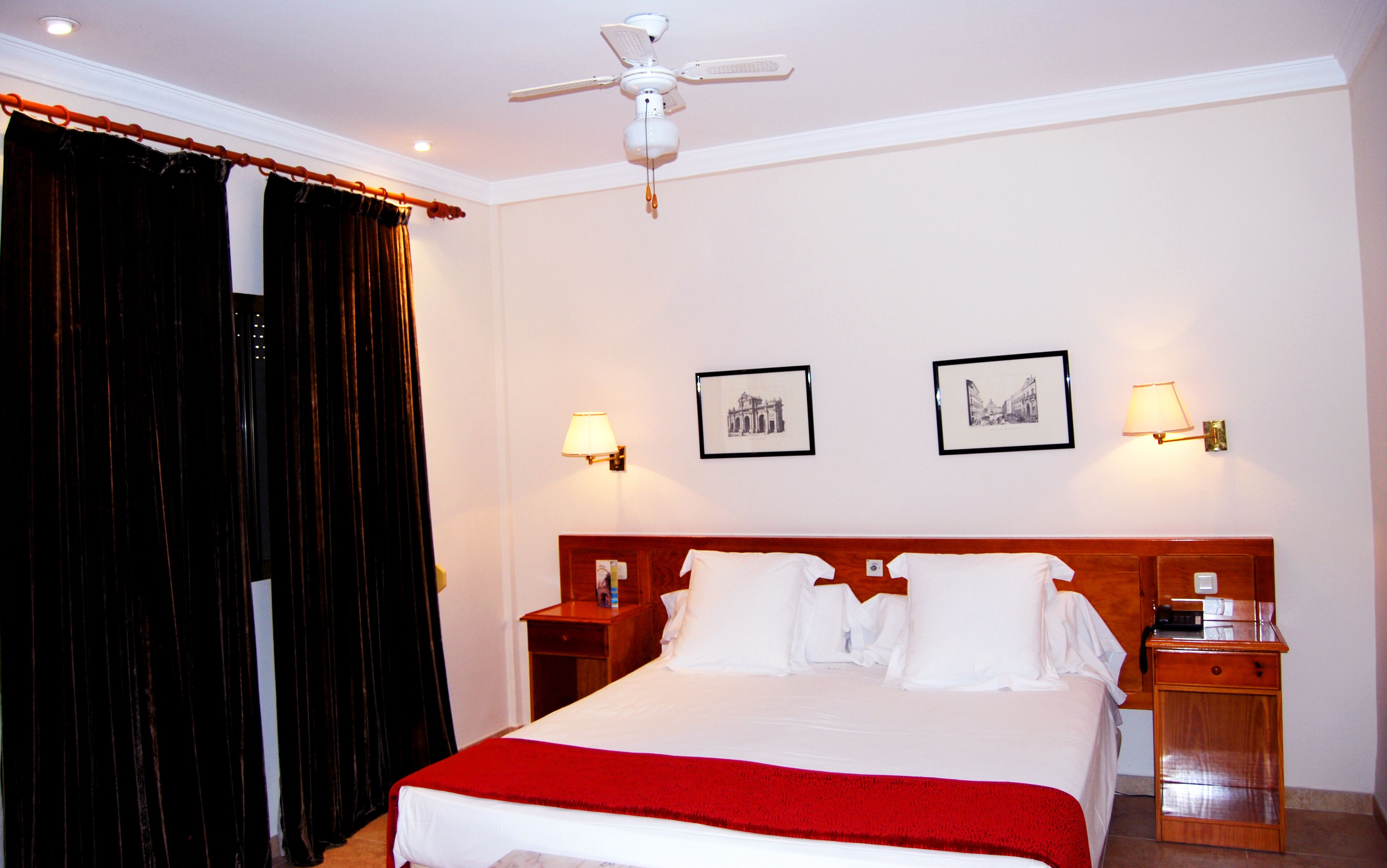 A hotel room with a bed, two bedside tables, a ceiling fan, black curtains, two wall-mounted lamps, and two framed pictures.