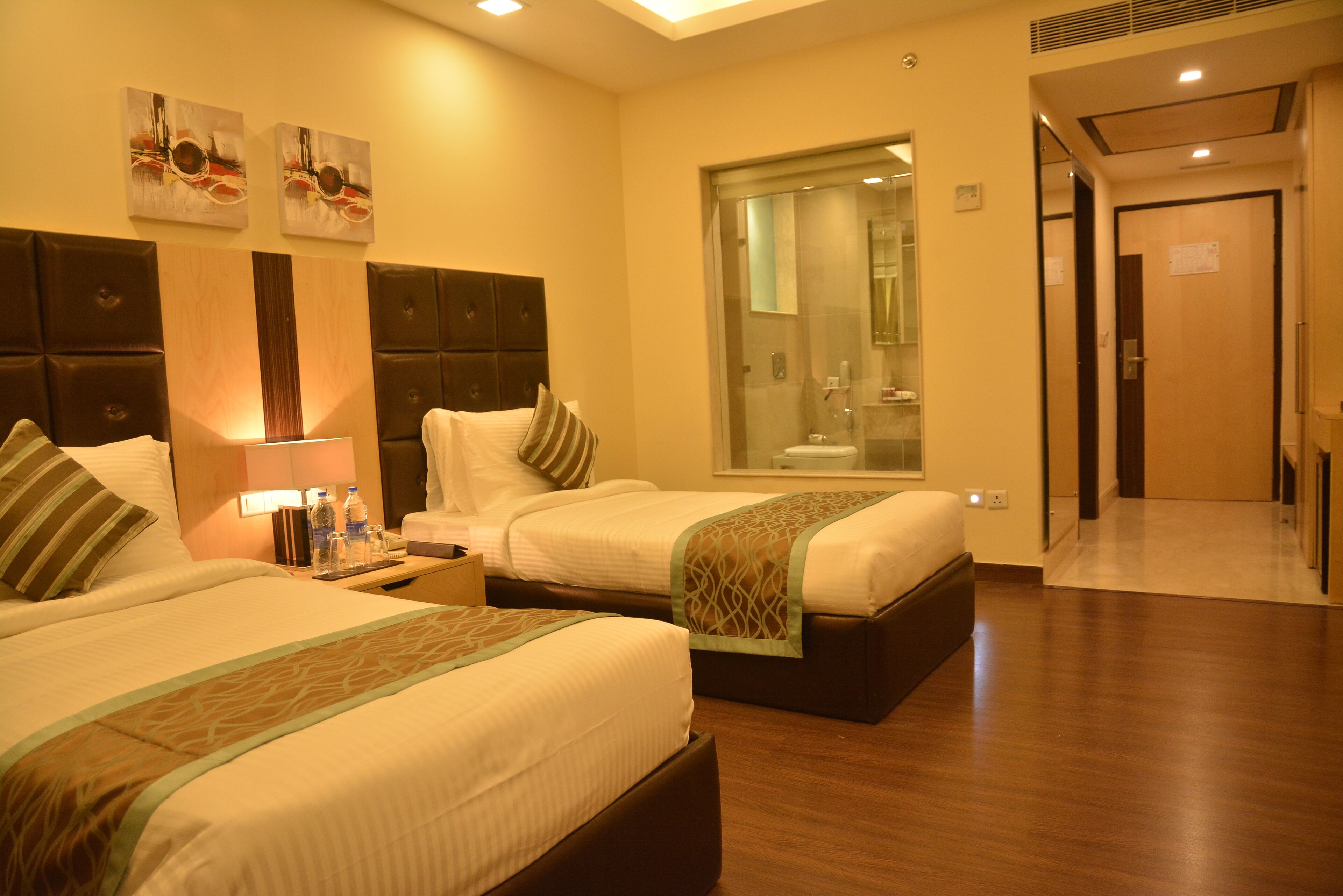 A hotel room with two beds, a bathroom with a glass shower, and a hallway leading to another room.