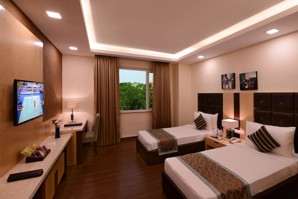 A hotel room with two beds, a TV, a desk with snacks, a window with curtains, and artwork on the wall.