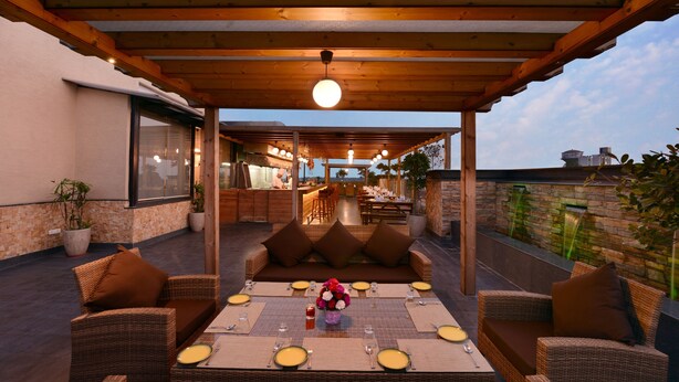 An outdoor patio with wicker furniture, a dining table set for four, and a bar area in the background. - Grand Plaza Jammu (Jammu)