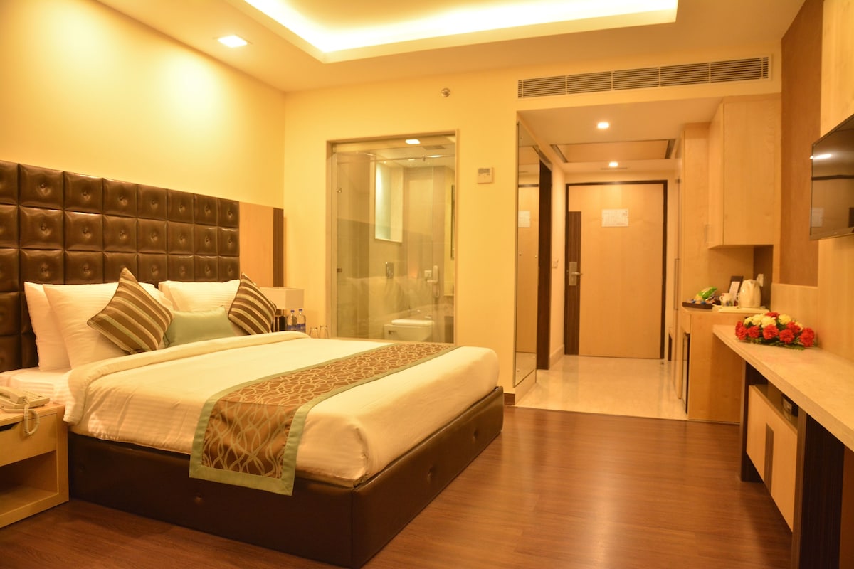 A hotel room with a large bed, a bedside table, a nightstand, a bathroom with a glass partition, and a kitchenette area.