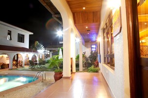 Uma área de piscina com piso ladrilhado, plantas em vasos e mobiliário de exterior.