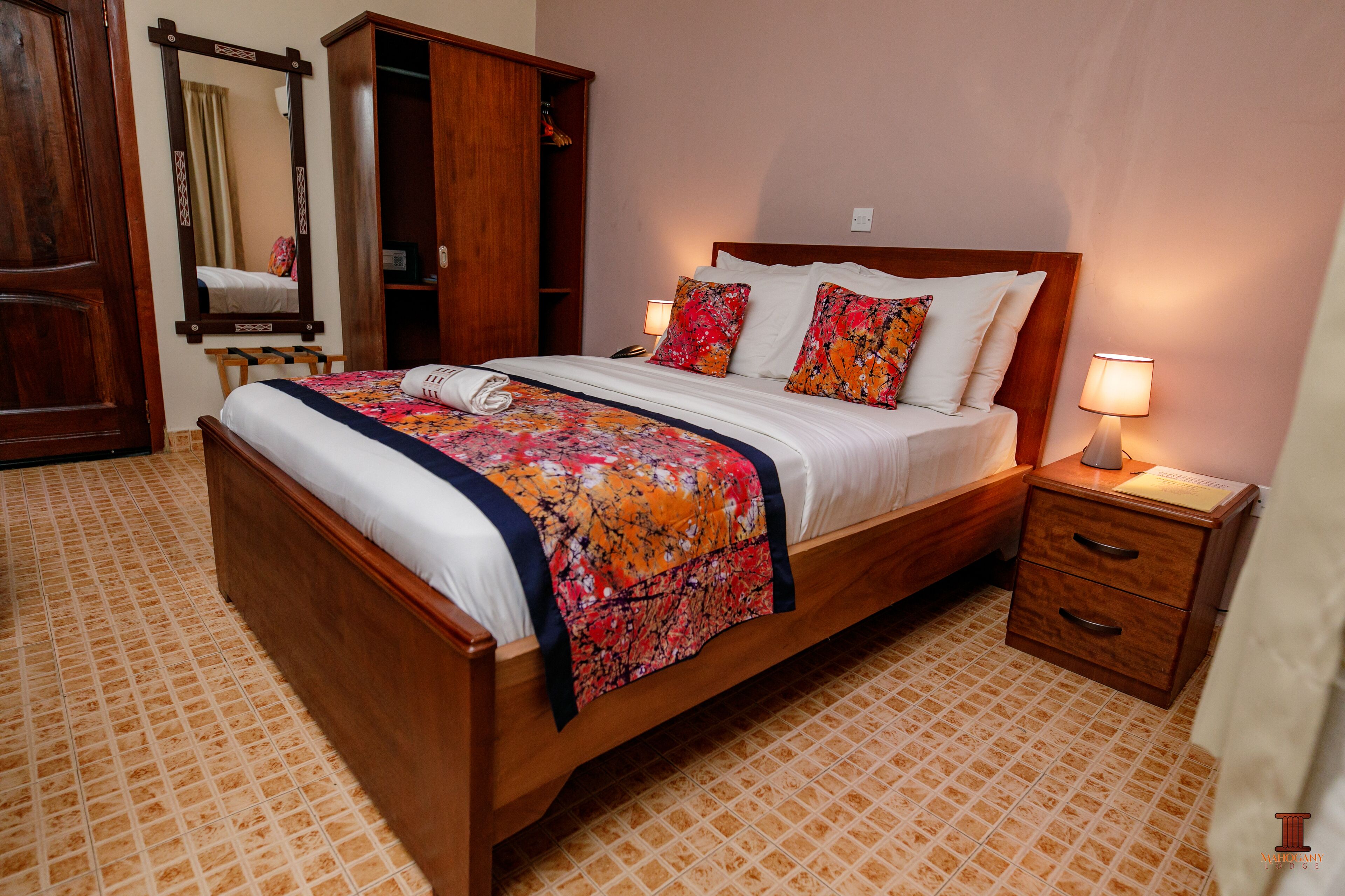 A neatly made bed with a floral bedspread, a wooden nightstand with a lamp, and a wardrobe with a mirror.
