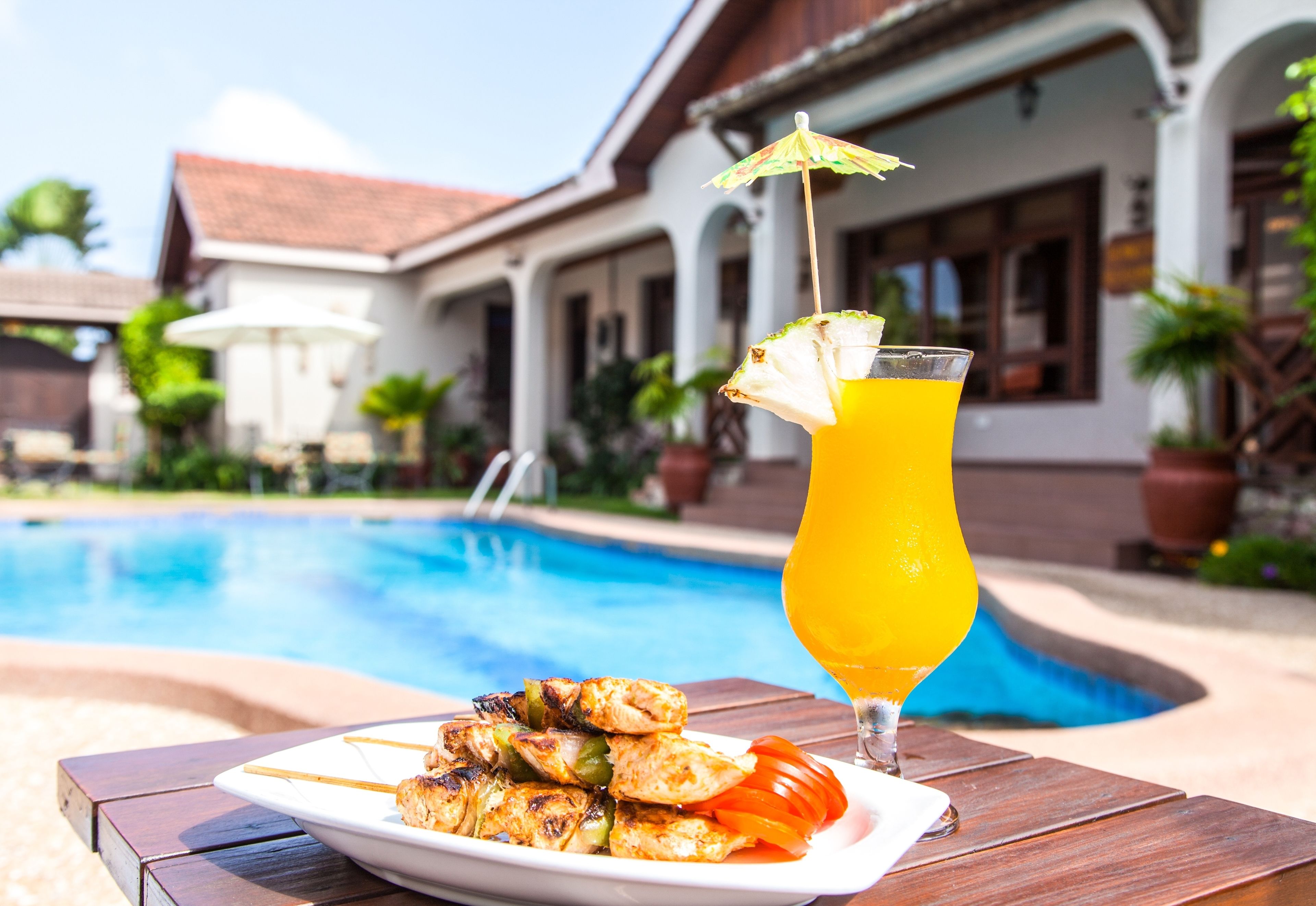 Una mesa junto a la piscina con brochetas de pollo a la parrilla y un vaso de jugo de naranja, con un edificio al fondo.
