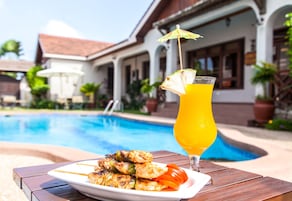 A poolside table with grilled chicken skewers and a glass of orange juice, with a building in the background.