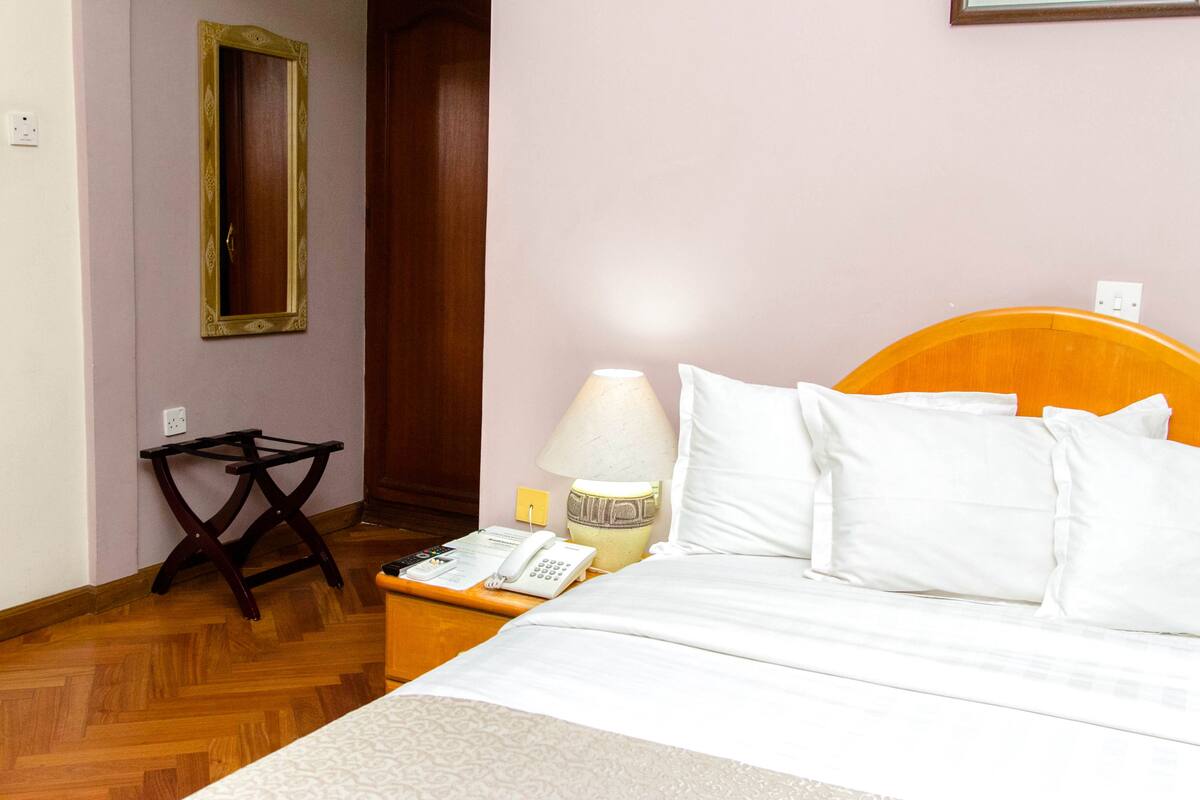A hotel room with a wooden bed, white pillows, a bedside table with a lamp, a telephone, and a mirror on the wall.