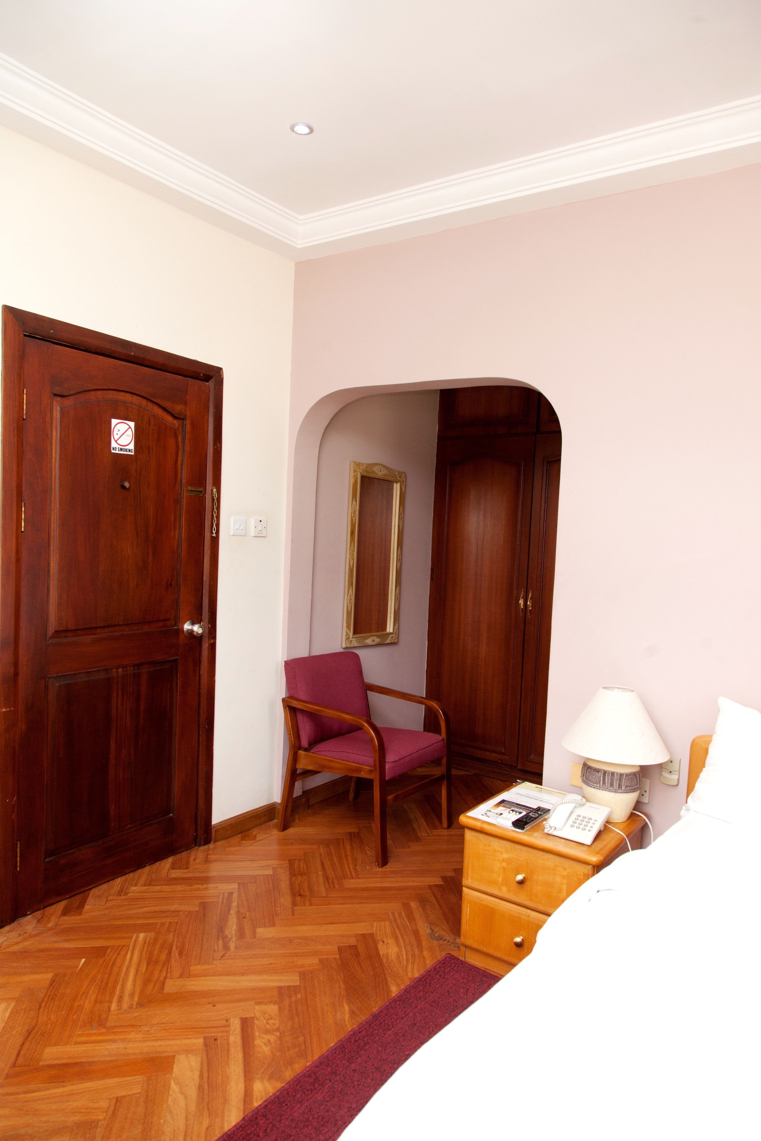 A bedroom with a wooden wardrobe, a chair, a bedside table with a lamp, and a bed.