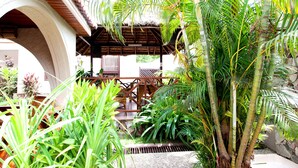 A covered outdoor area with a wooden bench and a thatched roof structure surrounded by lush greenery and palm trees. - Mahogany Lodge, Cantonments (Accra)