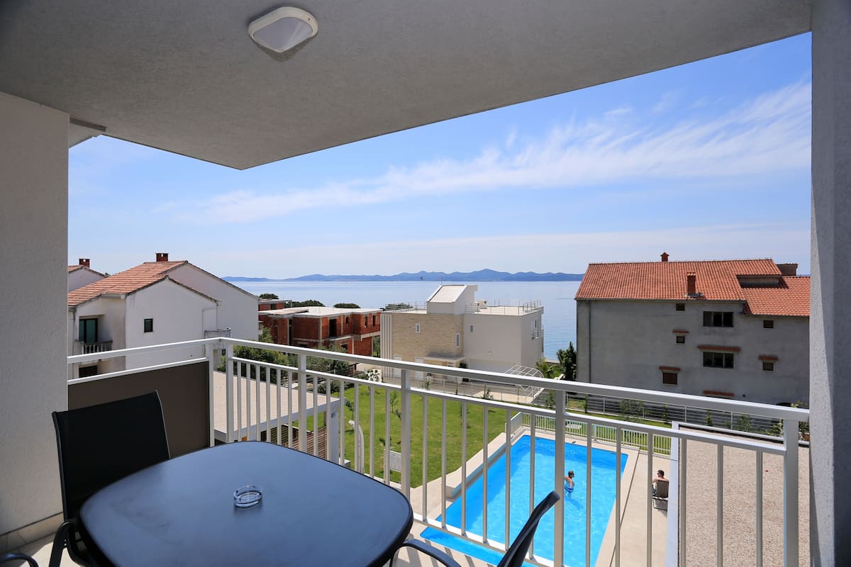 A balcony with a table and chairs, overlooking a swimming pool and a view of buildings and mountains.