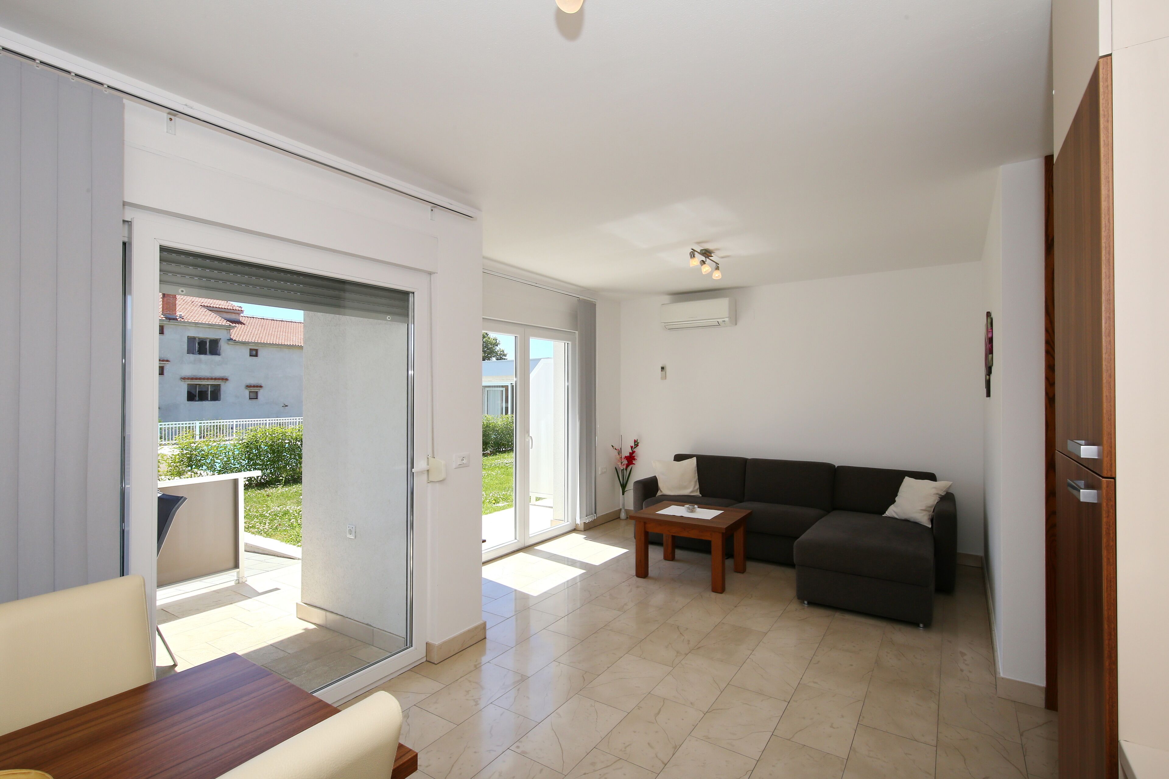 A modern living room with a sofa, a wooden coffee table, and a dining table. There is a sliding glass door leading to a balcony.
