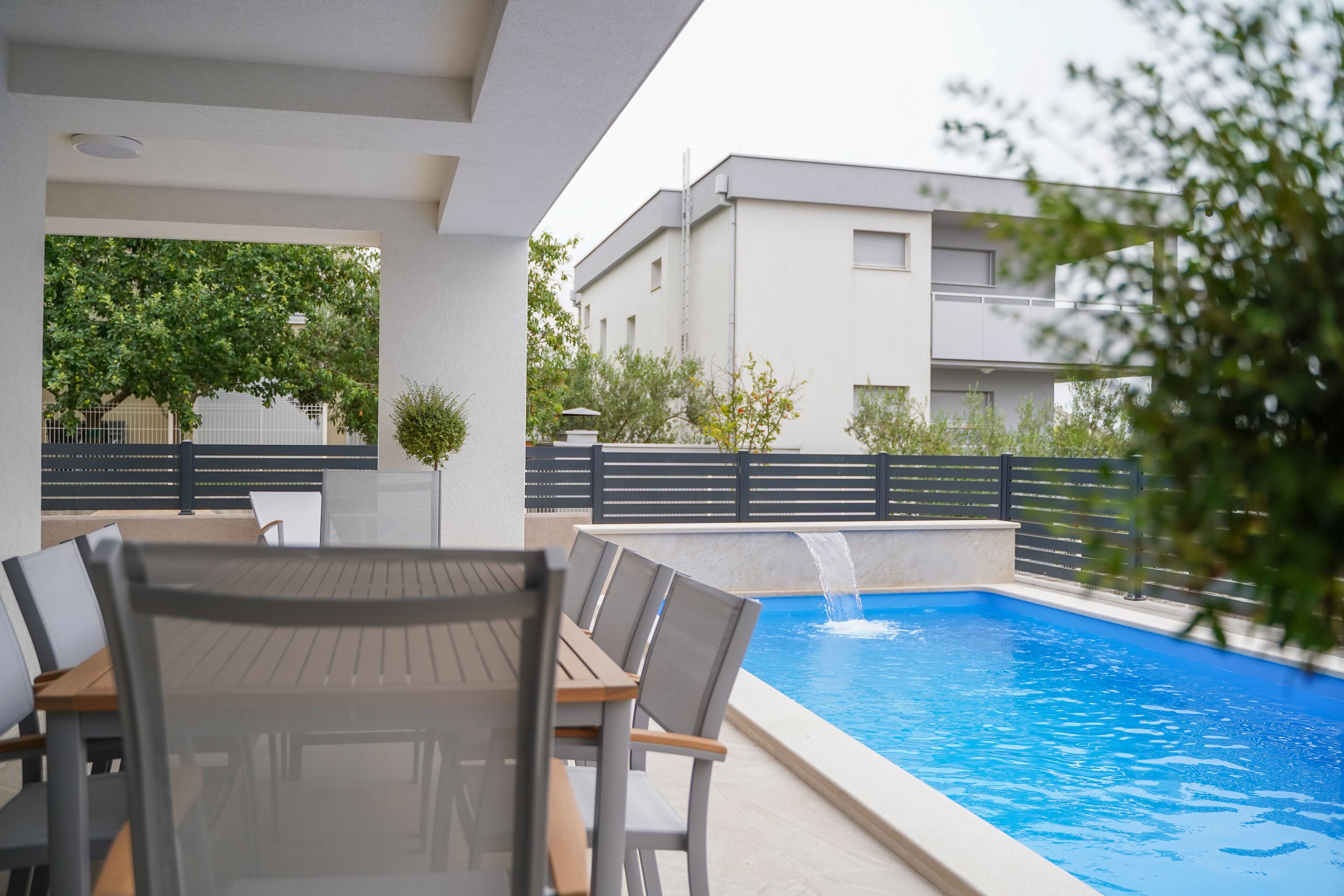 A poolside area with a table and chairs, a small waterfall feature, and a building in the background.