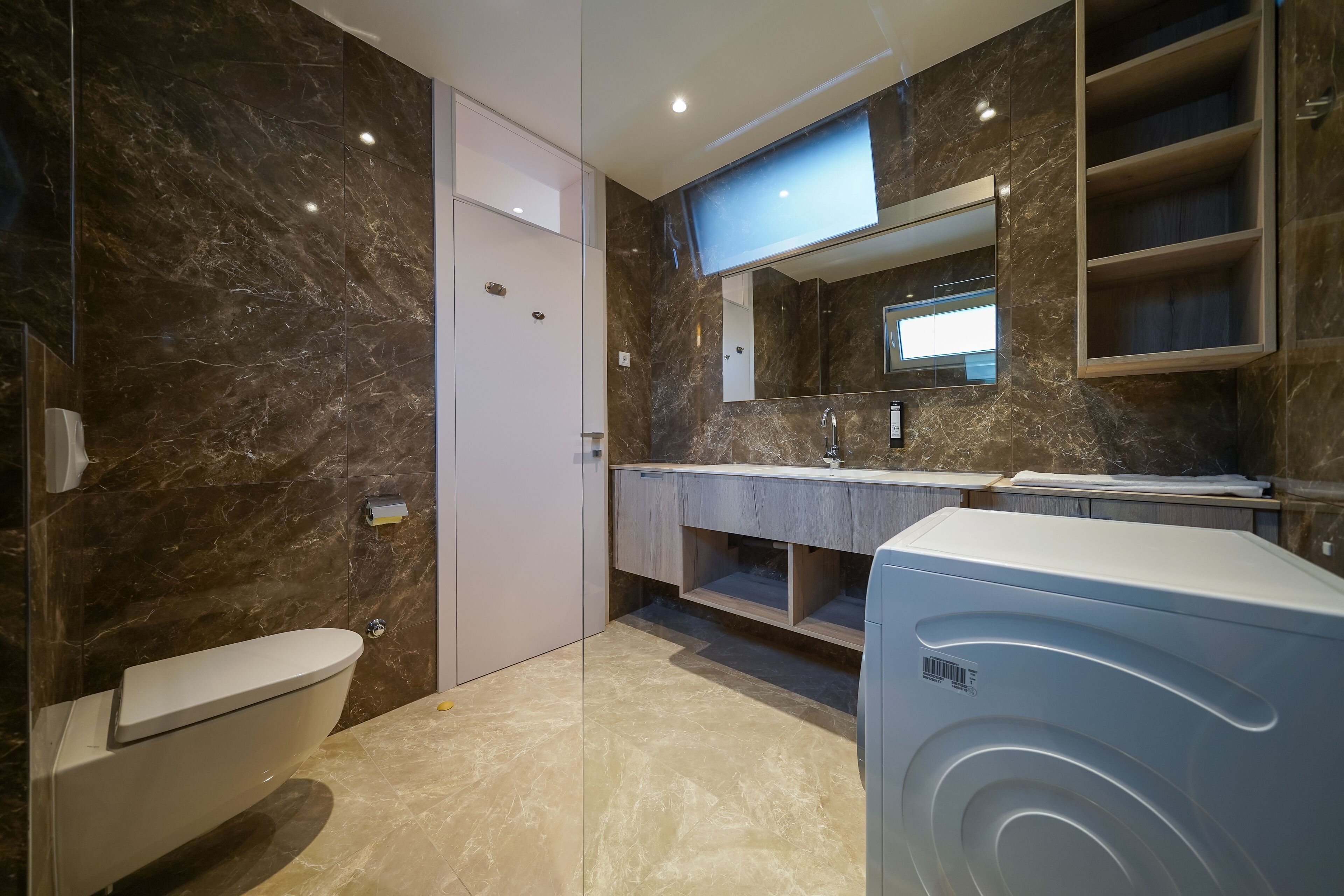 A modern bathroom with a marble wall, a built-in toilet, a vanity with a mirror, and a washing machine.