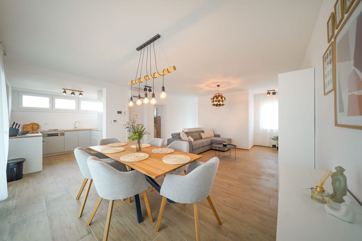 A modern kitchen with a dining area, featuring a wooden table with grey chairs, a hanging light fixture, and a grey sofa in the background.