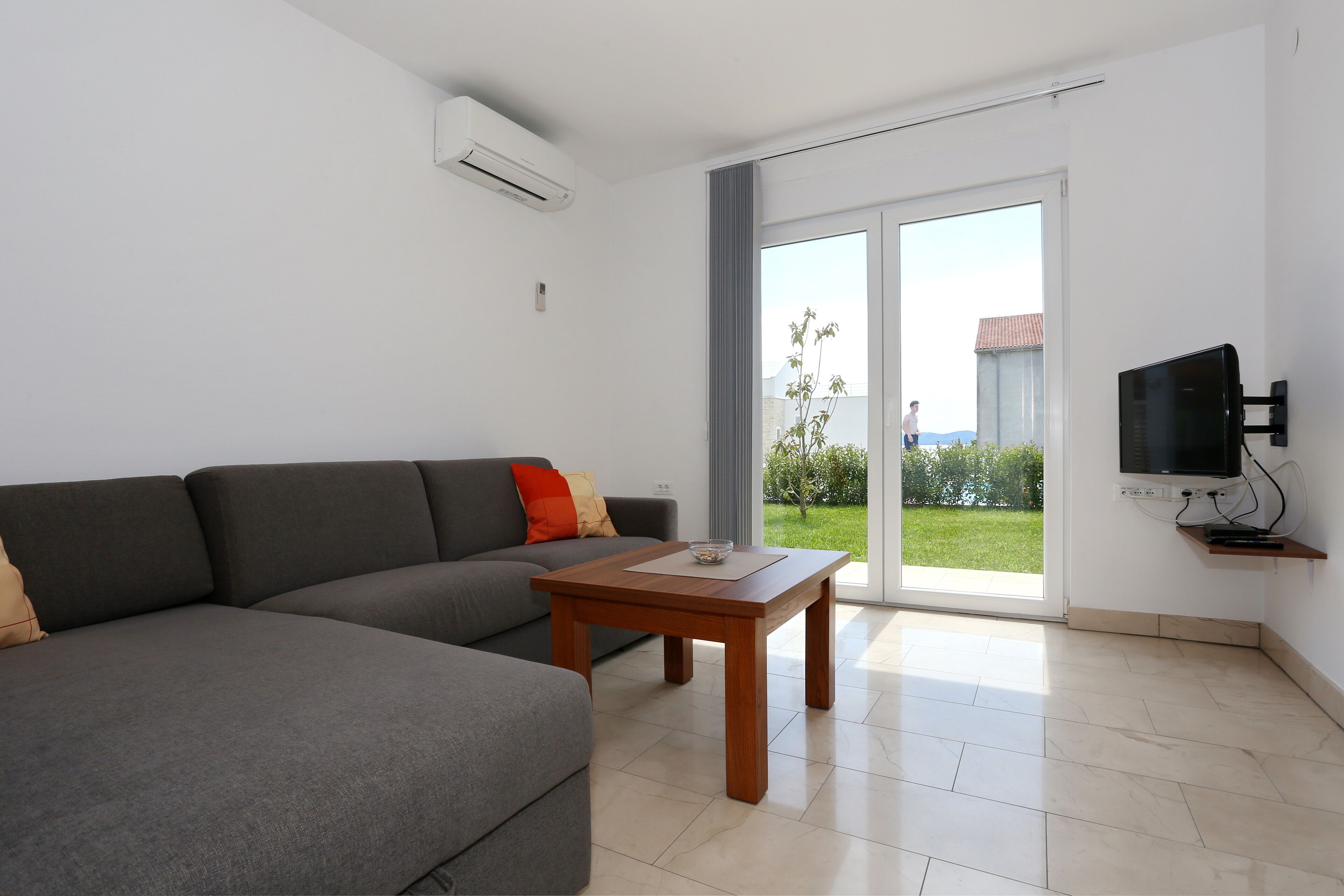 A modern living room with a grey sectional sofa, a wooden coffee table, a flat-screen TV mounted on the wall, and a sliding glass door leading to a balcony.