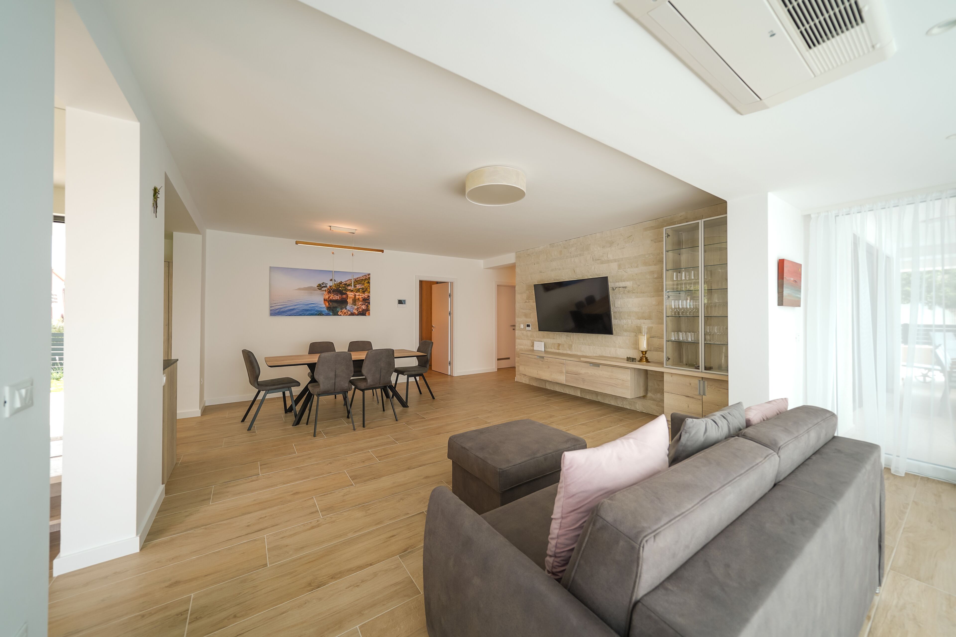A modern living room with a grey sofa, a dining table with chairs, a flat-screen TV mounted on a wooden cabinet, and a large window with sheer curtains.