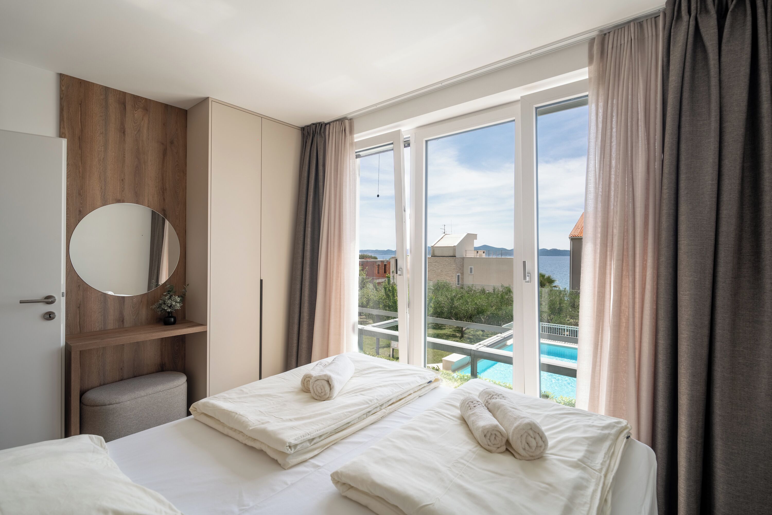 A hotel room with a bed, a round mirror, a small stool, and a view of a pool and buildings.