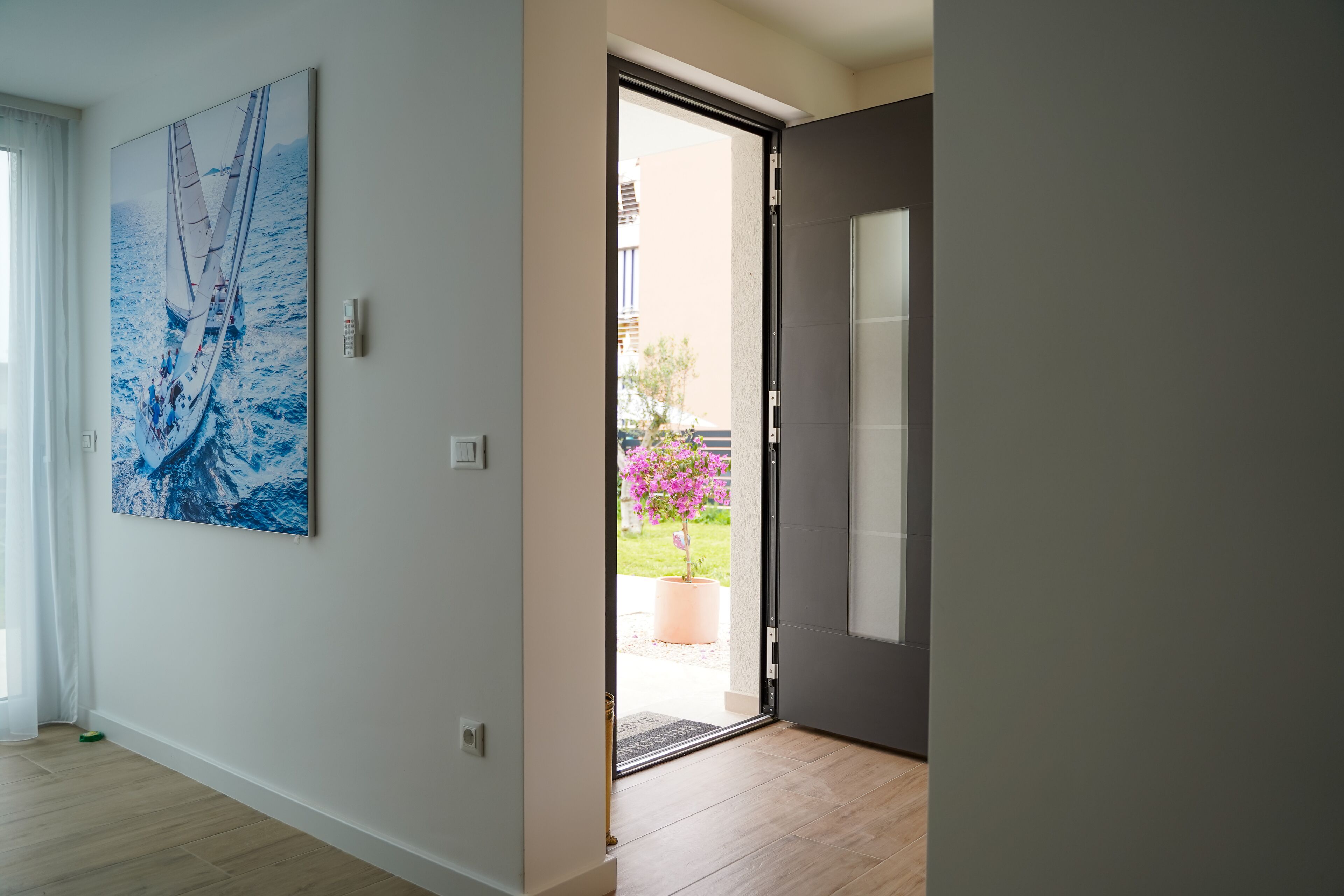 A hallway with a large sailboat painting on the wall, a door leading outside, and a potted plant with pink flowers.