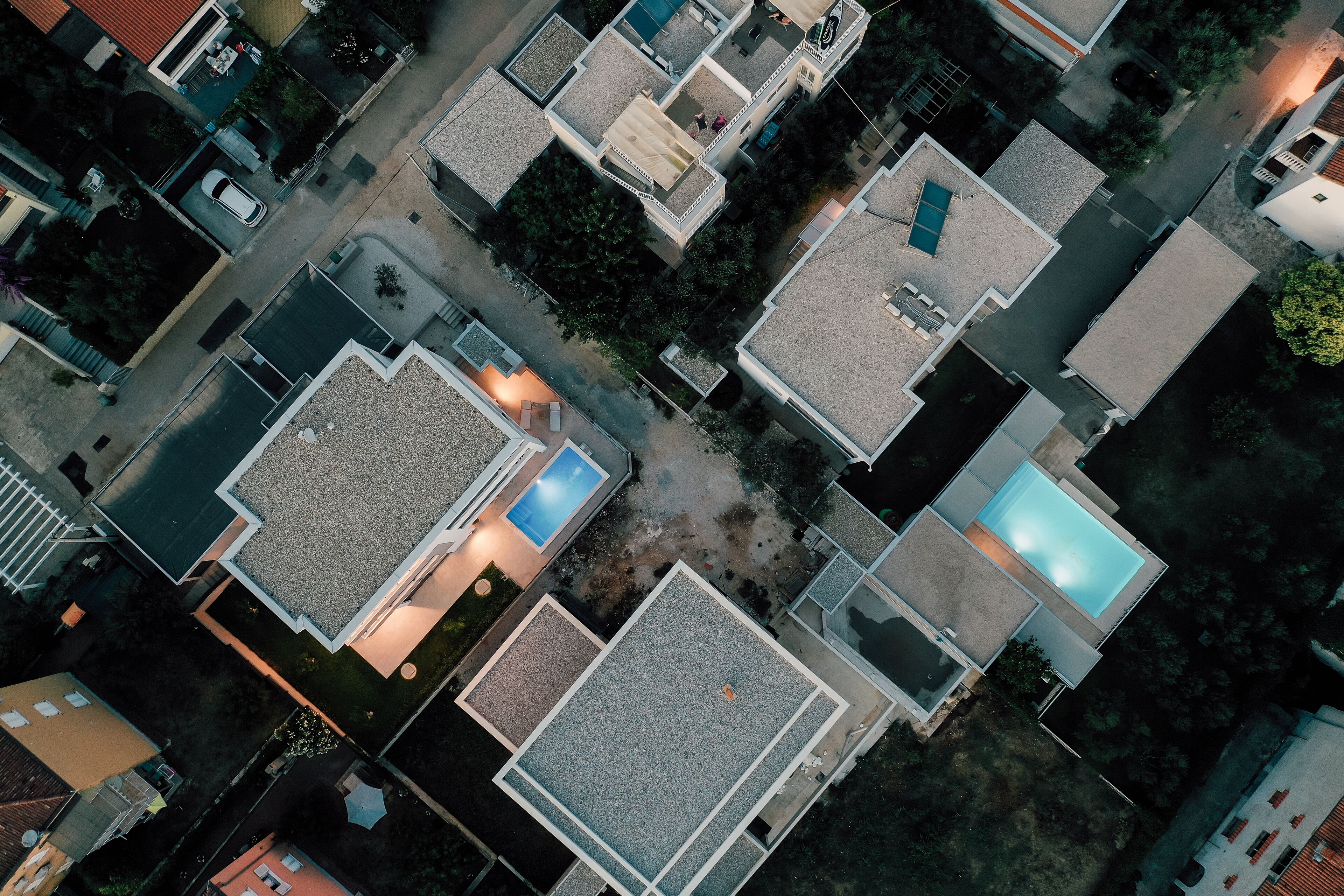 An aerial view of a residential area with multiple houses, each featuring a distinct roof design and a visible swimming pool.