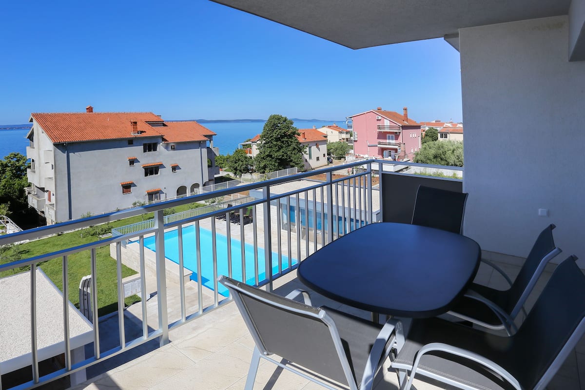 A balcony with a table and chairs overlooking a swimming pool and a coastal view.