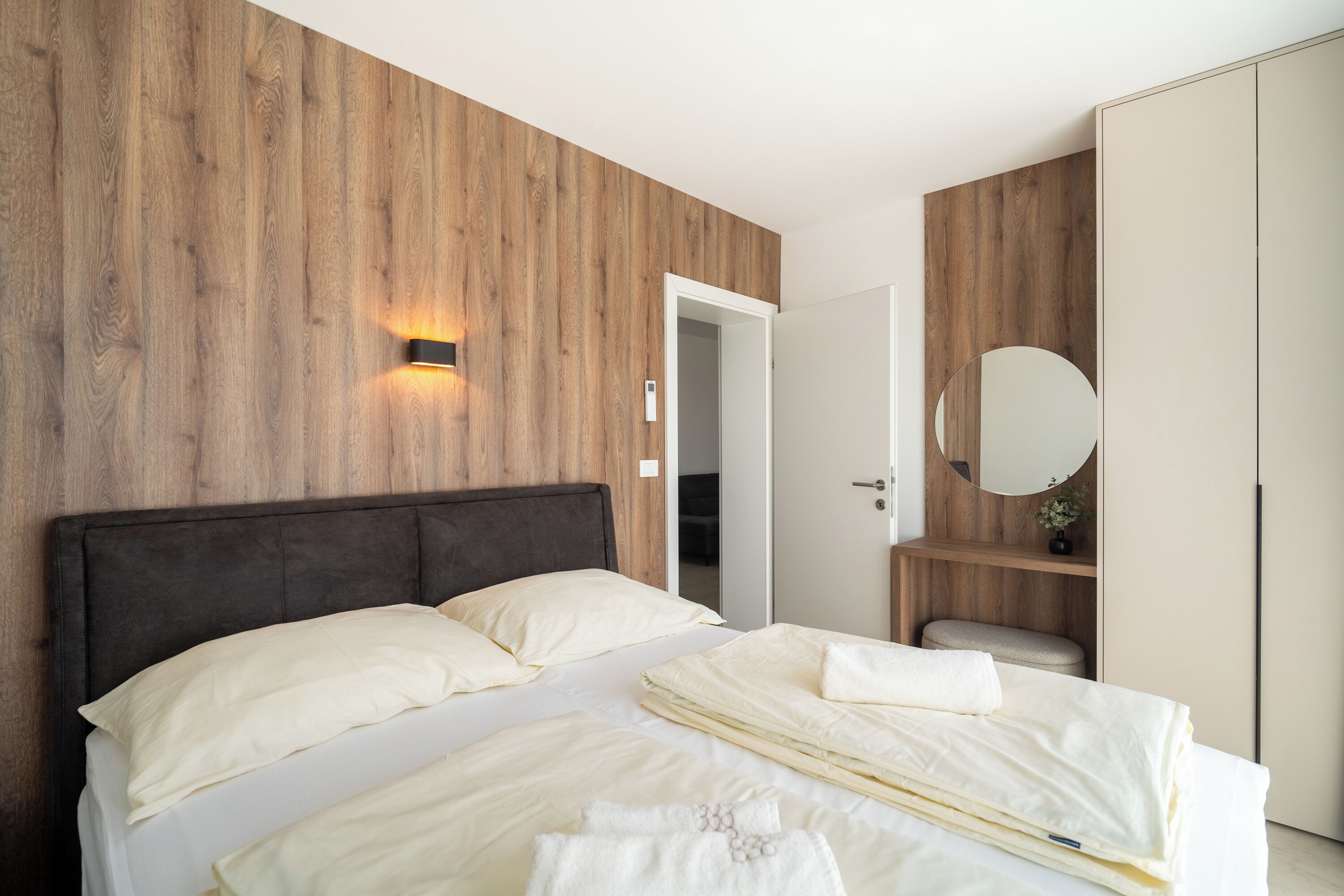 A modern bedroom with a wooden wall panel, a bed with white bedding, a bedside table with a mirror, and a wardrobe.