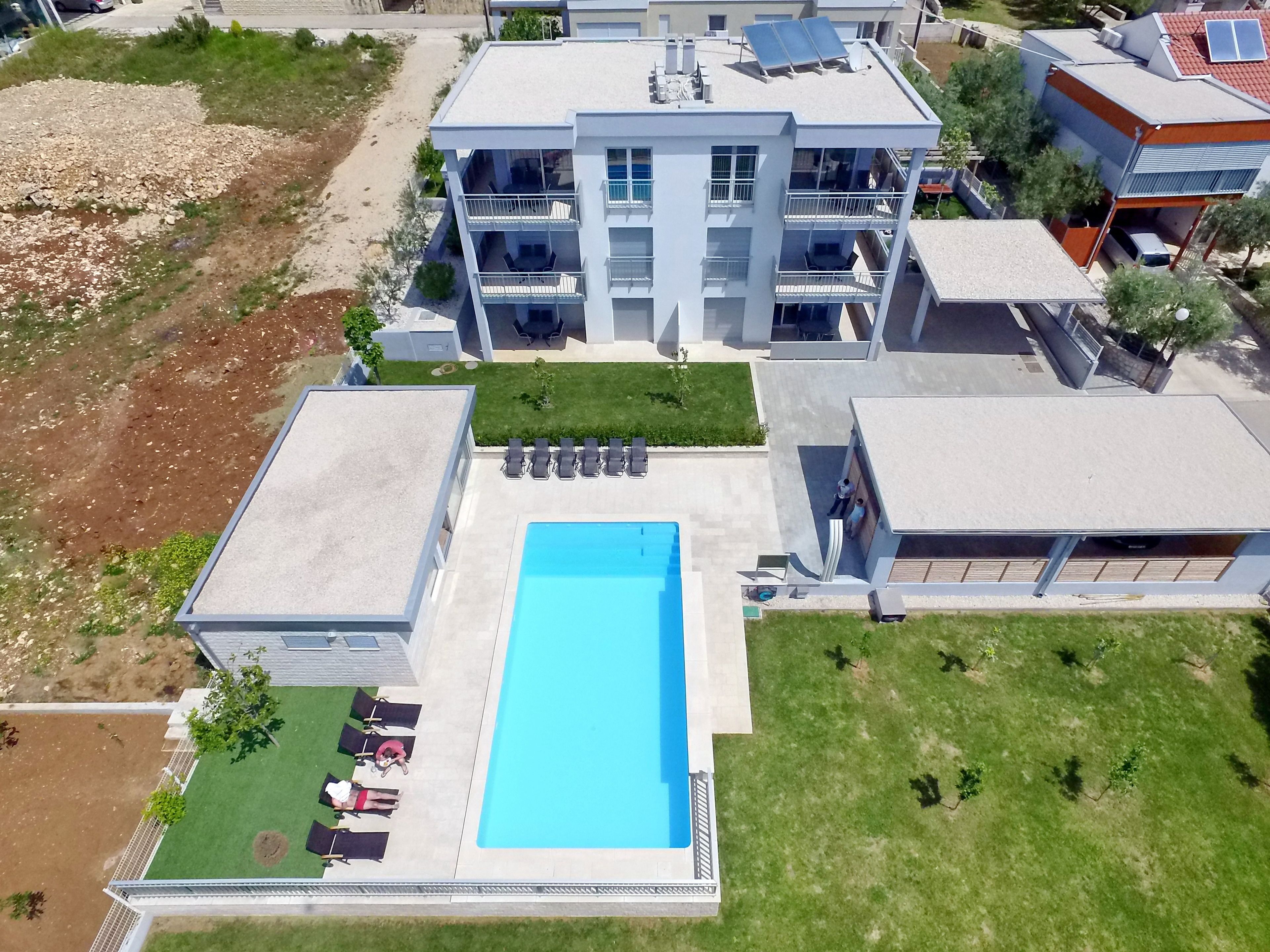 A modern multi-story building with a central swimming pool, surrounded by a well-maintained lawn and a few lounge chairs.