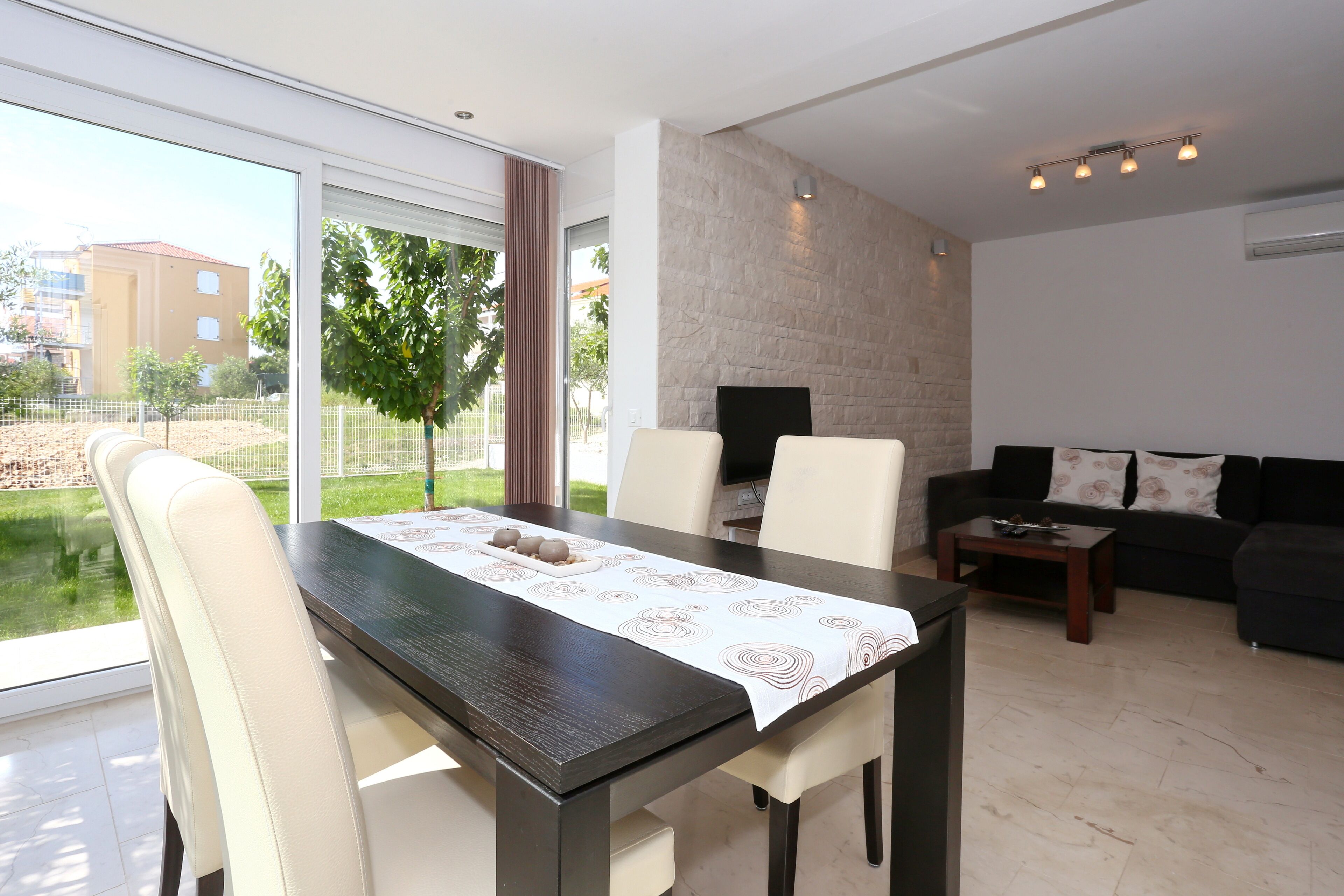A modern living room with a dining area, featuring a large window, a stone accent wall, and a black sofa.