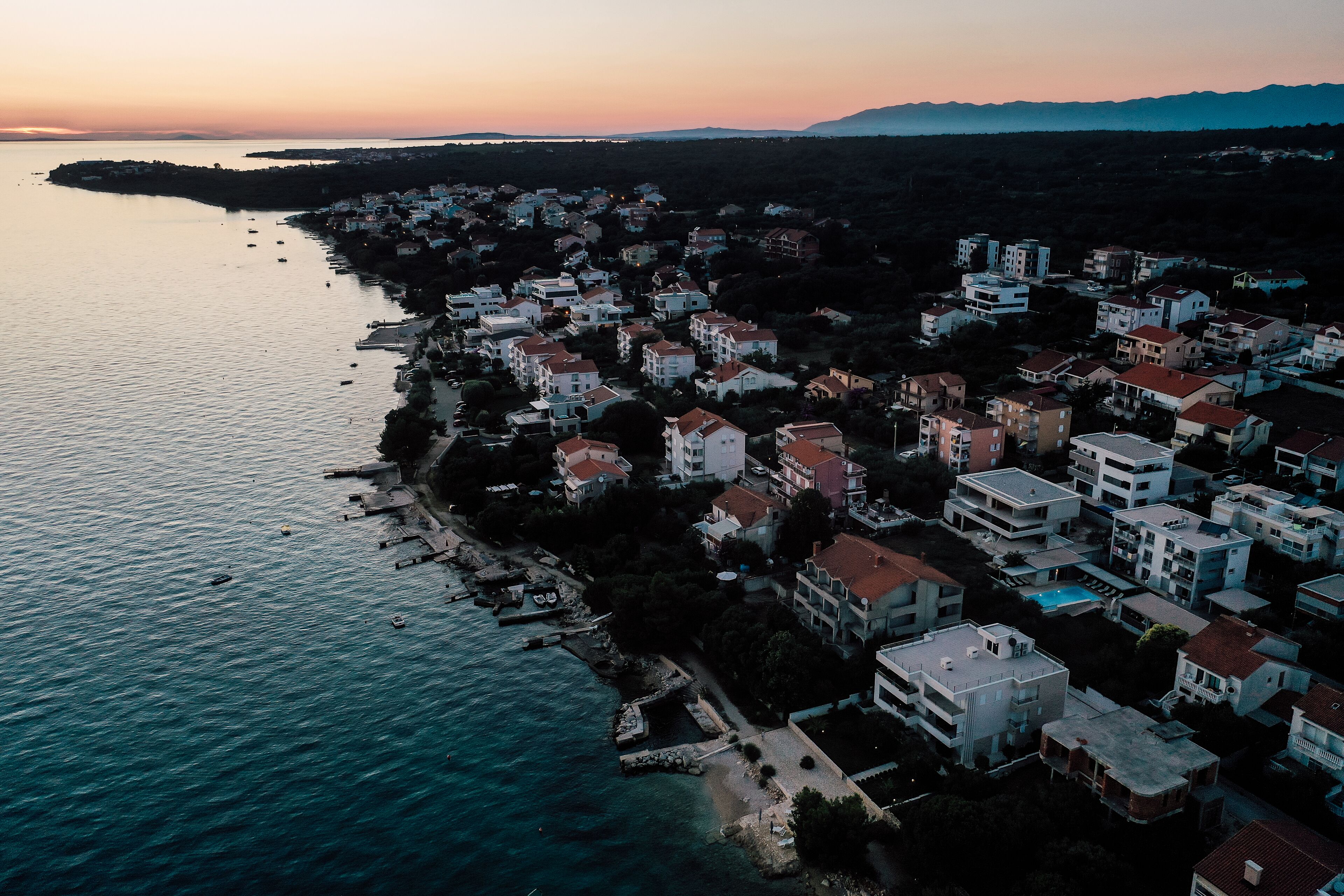 An aerial view of a coastal residential area with buildings along the shoreline, a clear sky, and a calm sea.