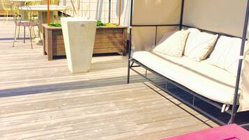 Una terraza de madera con un área de estar techada, una mesa con sillas y plantas en macetas.