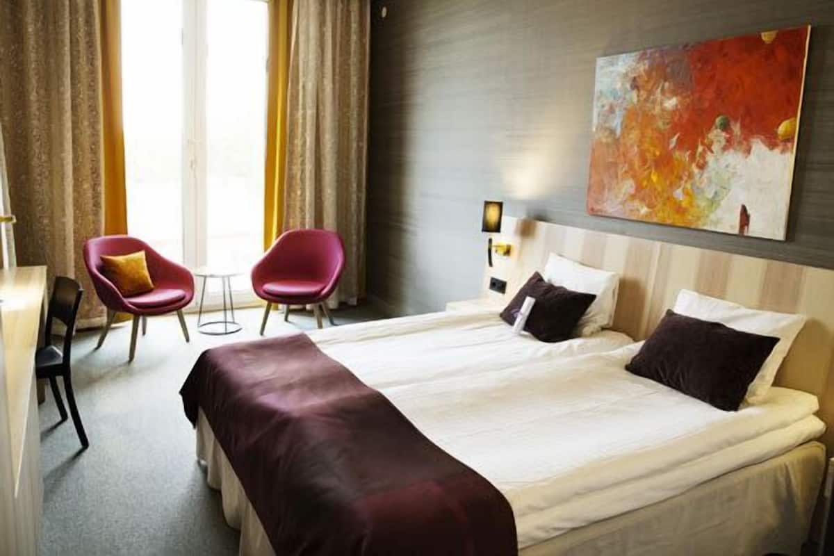 A hotel room with a large bed, two red chairs, a small table, and a painting on the wall.