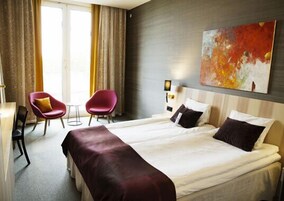 Habitación de hotel con una cama grande, dos sillas rojas, una mesita y un cuadro en la pared.