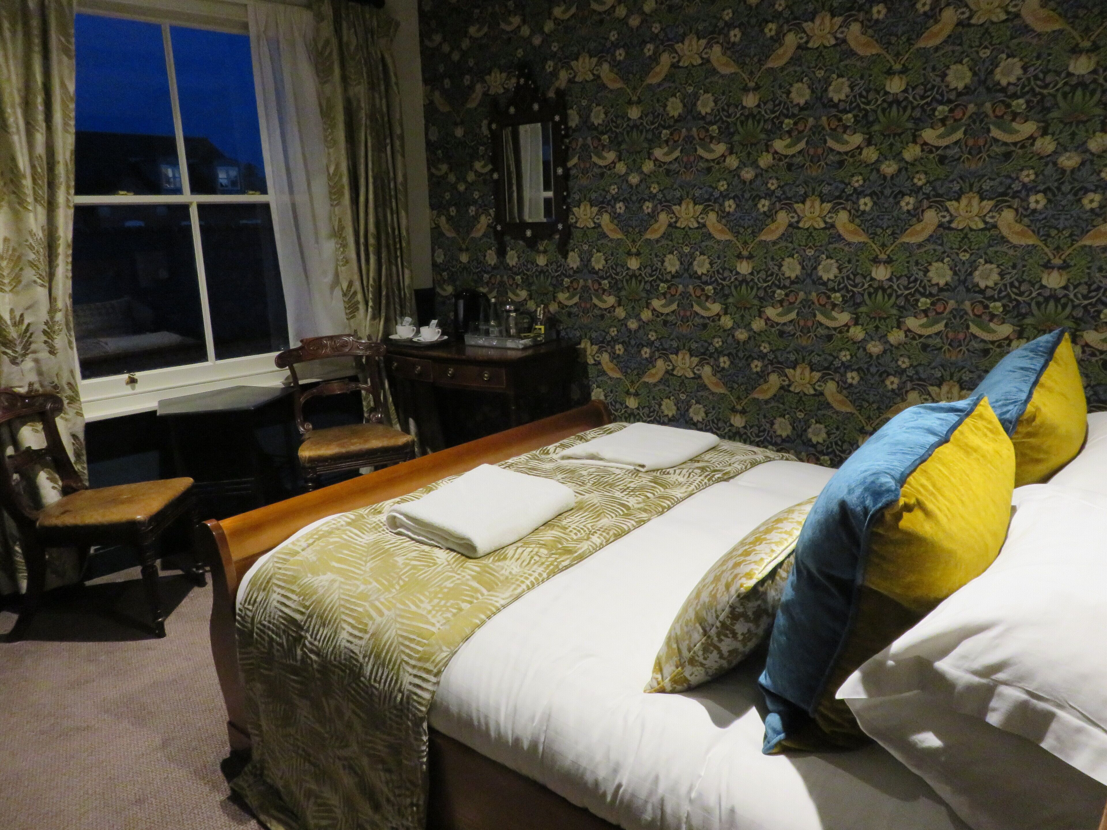 A neatly made bed with a patterned bedspread and two pillows, a wooden nightstand with a lamp, a chair, and a desk with a cup and saucer.