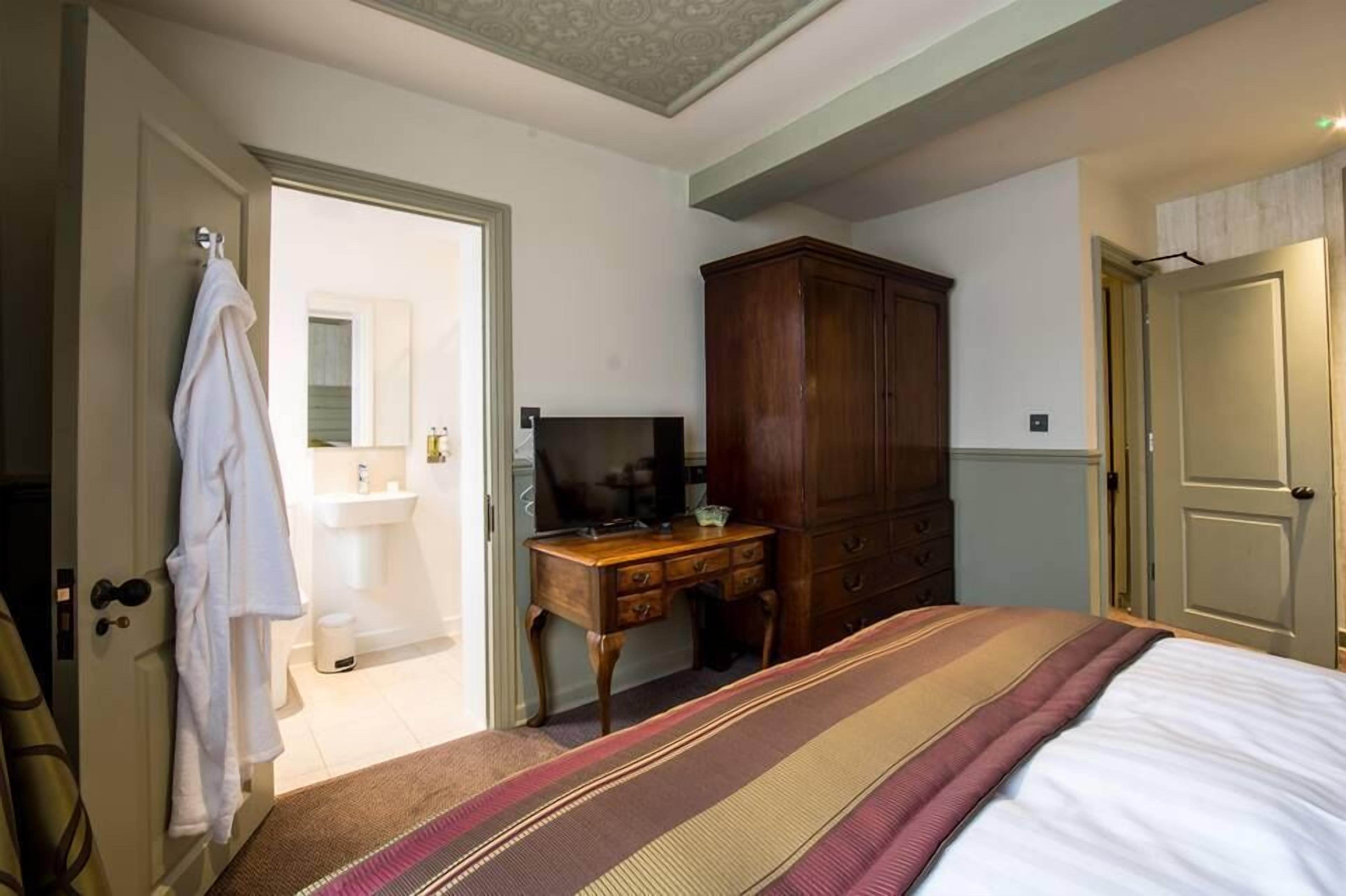 Un dormitorio con una cama, un armario de madera, un escritorio con televisor, y un baño visible a través de una puerta abierta.