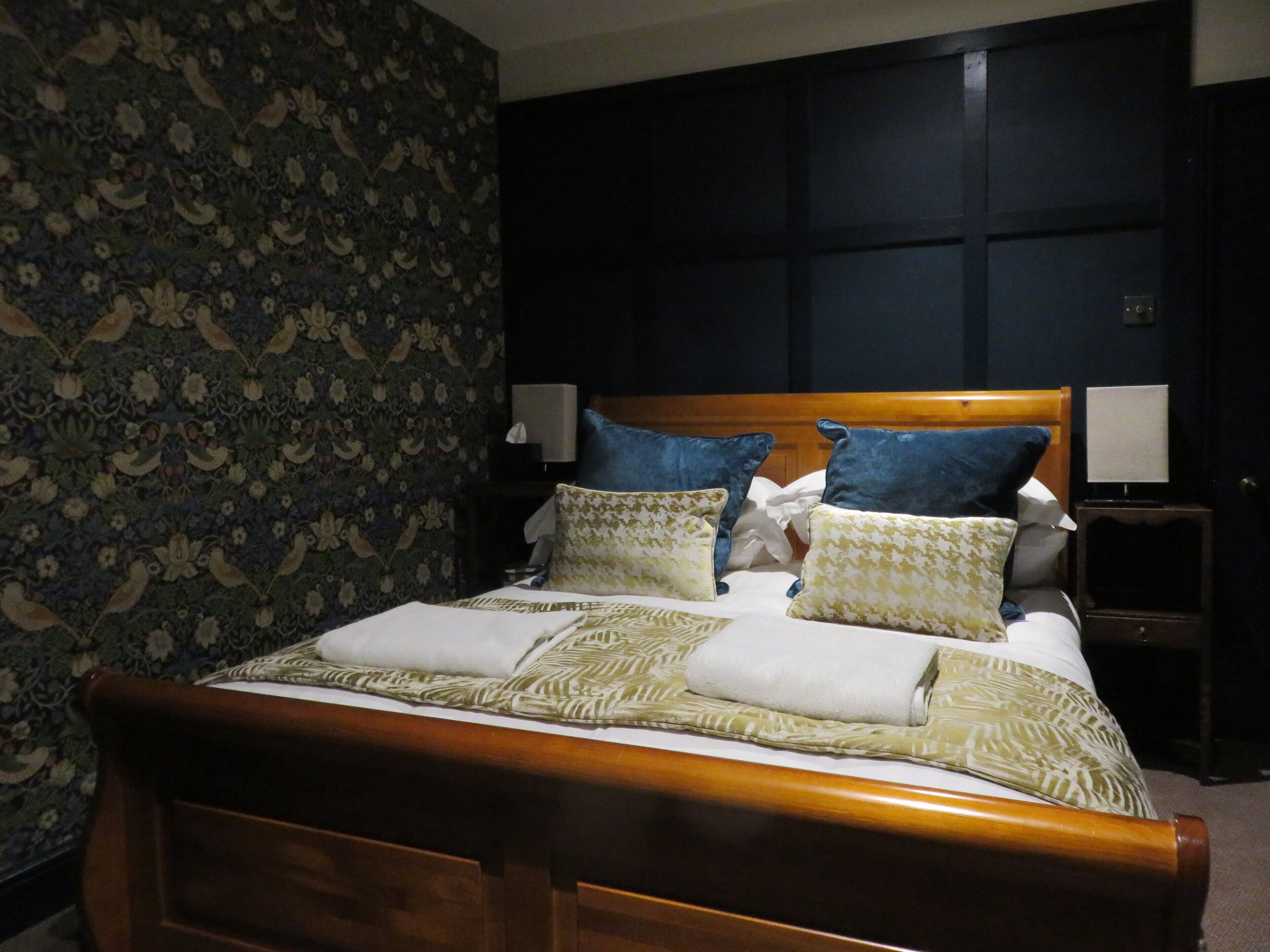 Uma cama bem arrumada com cabeceira de madeira, ladeada por duas mesas de cabeceira com abajures, em um quarto com papel de parede floral e painéis escuros nas paredes.