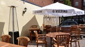 An outdoor seating area with wooden tables and chairs, a white umbrella with 'VELTINS' branding, and a tall, narrow lamp post. - The Cardinals Hat (Worcester)