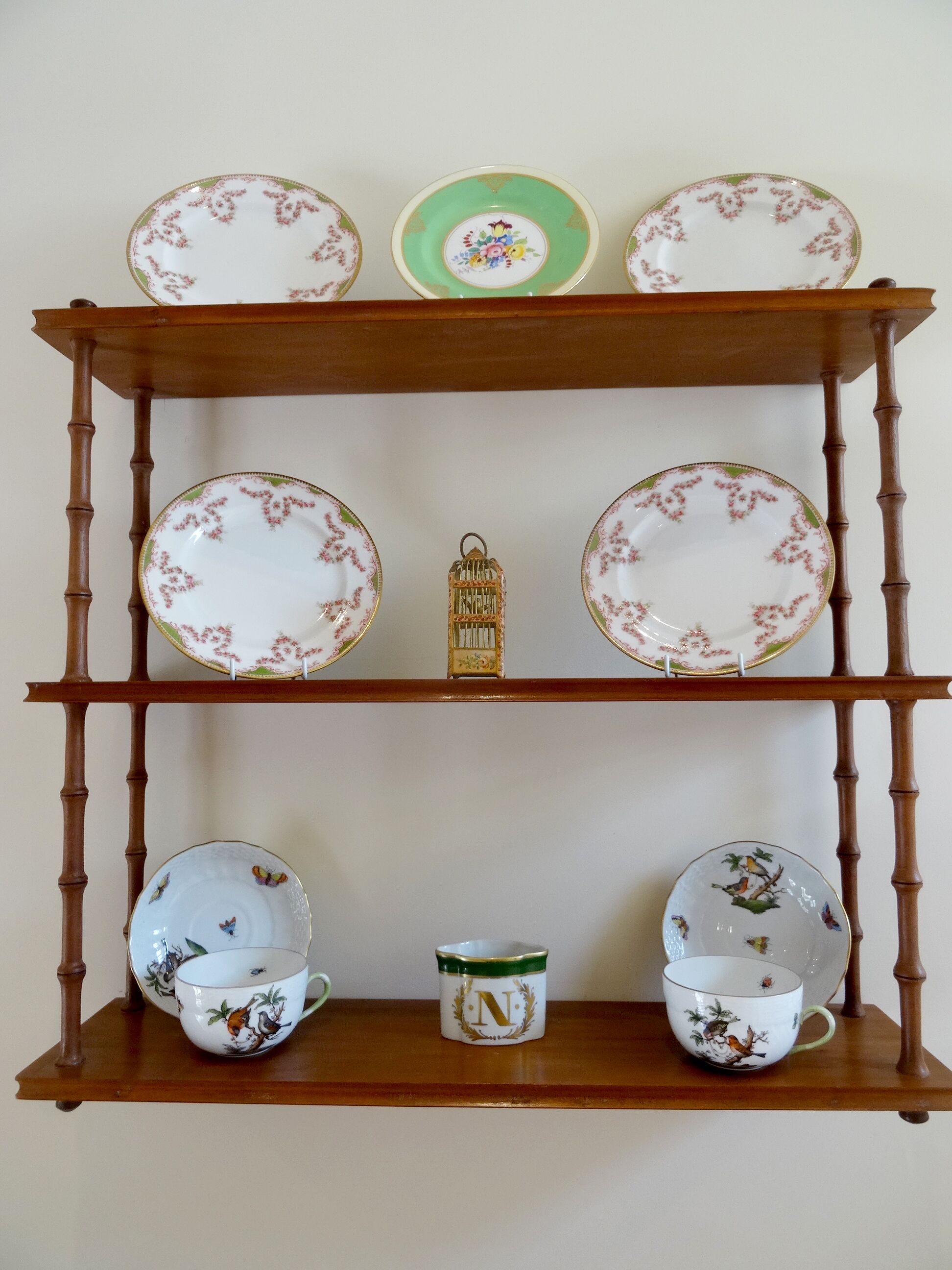 A wooden three-tiered shelf with decorative plates, a birdcage, and a teacup with a bird design.