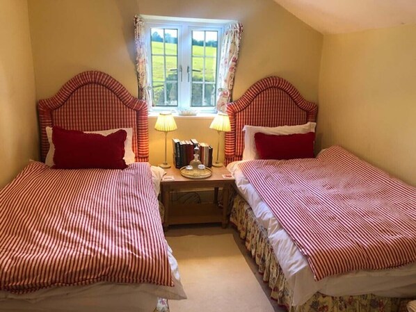 A room with two beds, each with a red and white striped bedspread, a small table with books and a lamp, and a window with a view of a green field. - Hollamoor Farm B&B (Barnstaple)
