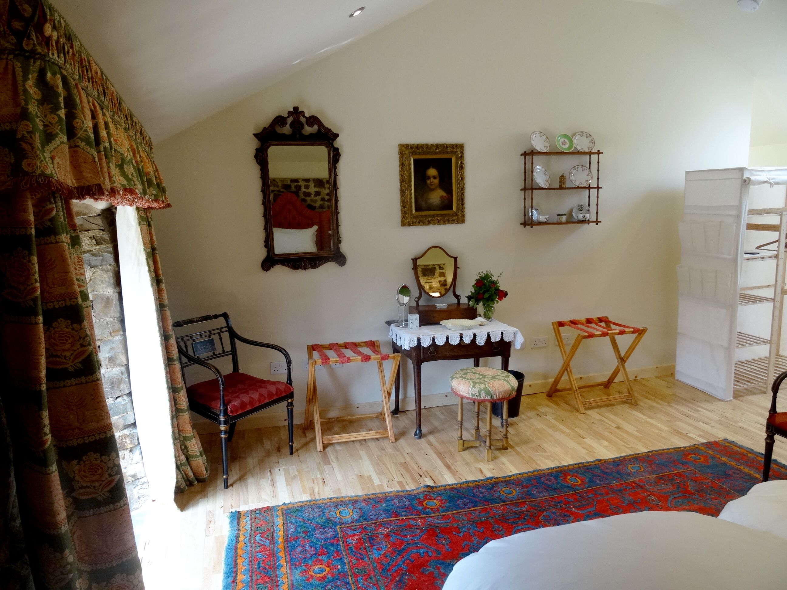 A room with a bed, a chair, a small table with a mirror, a shelf with decorative items, and a patterned rug.