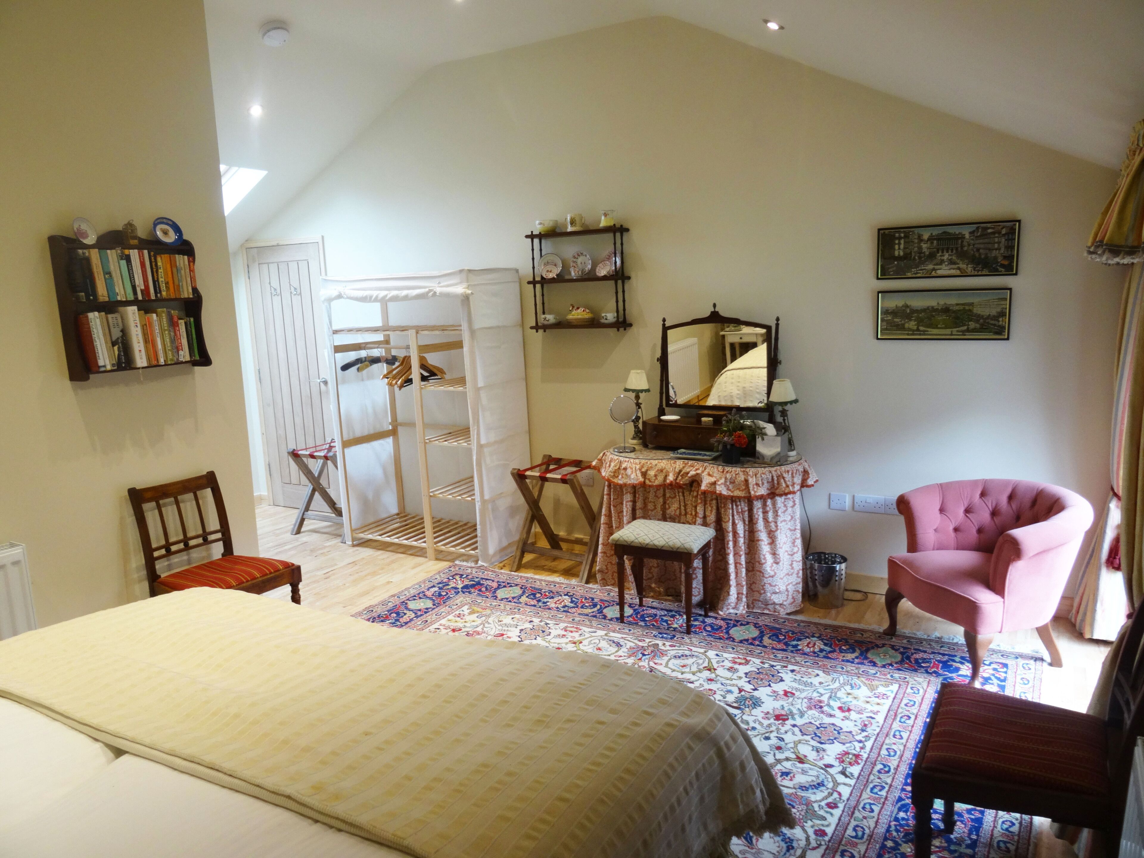 Un dormitorio con cama, una silla, tocador con espejo, estantería y alfombra estampada.