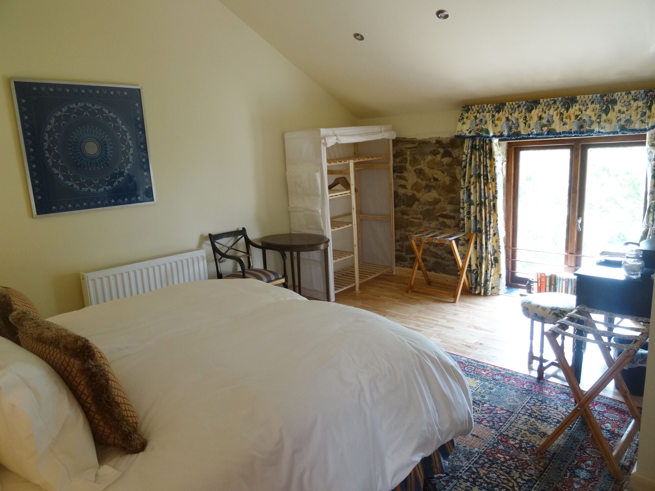 A bedroom with a large bed, a chair, a small table, a wardrobe, and a window with curtains.